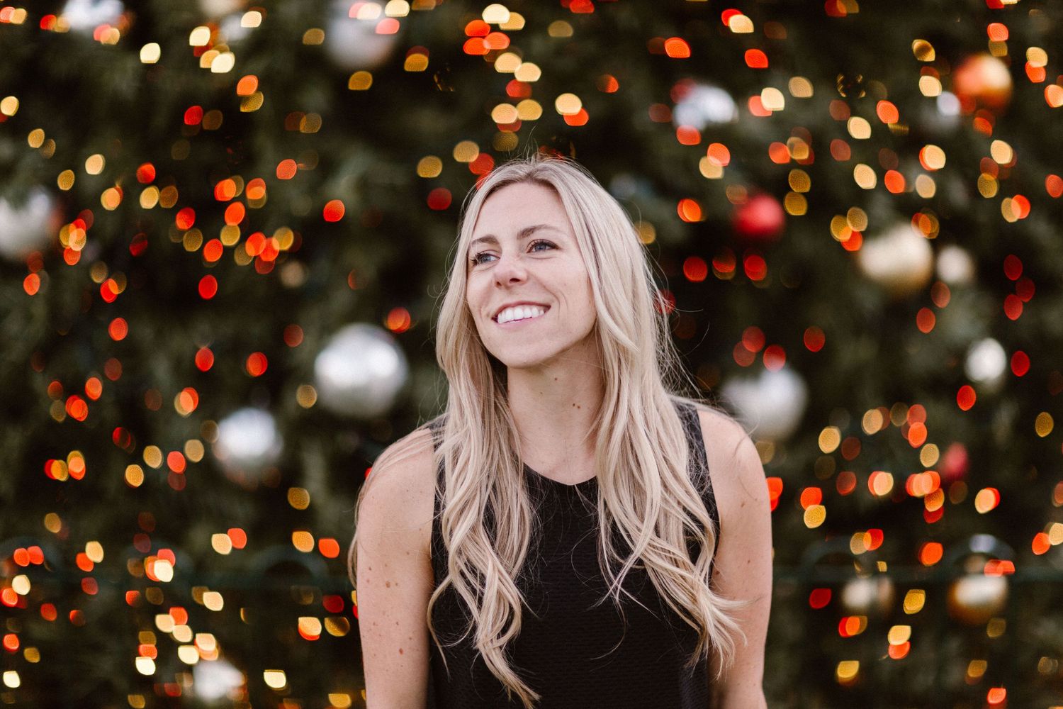 Smiling blonde woman in black top posing in front of a decorated Christmas tree with colorful bokeh lights.