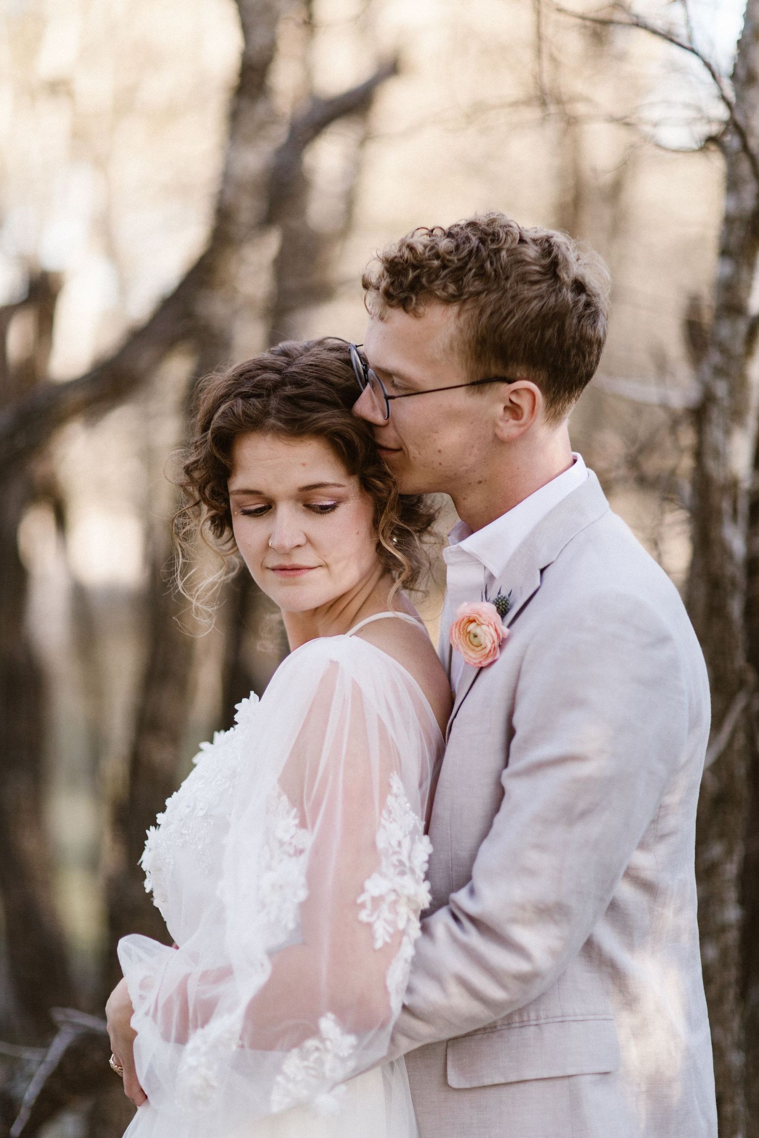 Groom in gray suit kissing bride's head while embracing her in sheer lace wedding dress outdoors.