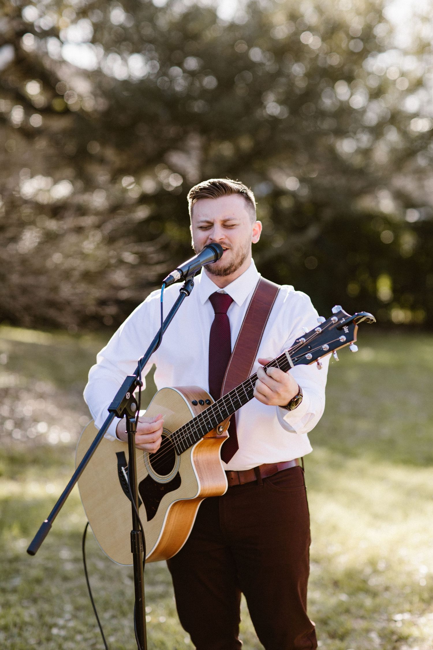 Male musician in white shirt and burgundy tie plays acoustic guitar and sings at outdoor wedding ceremony.