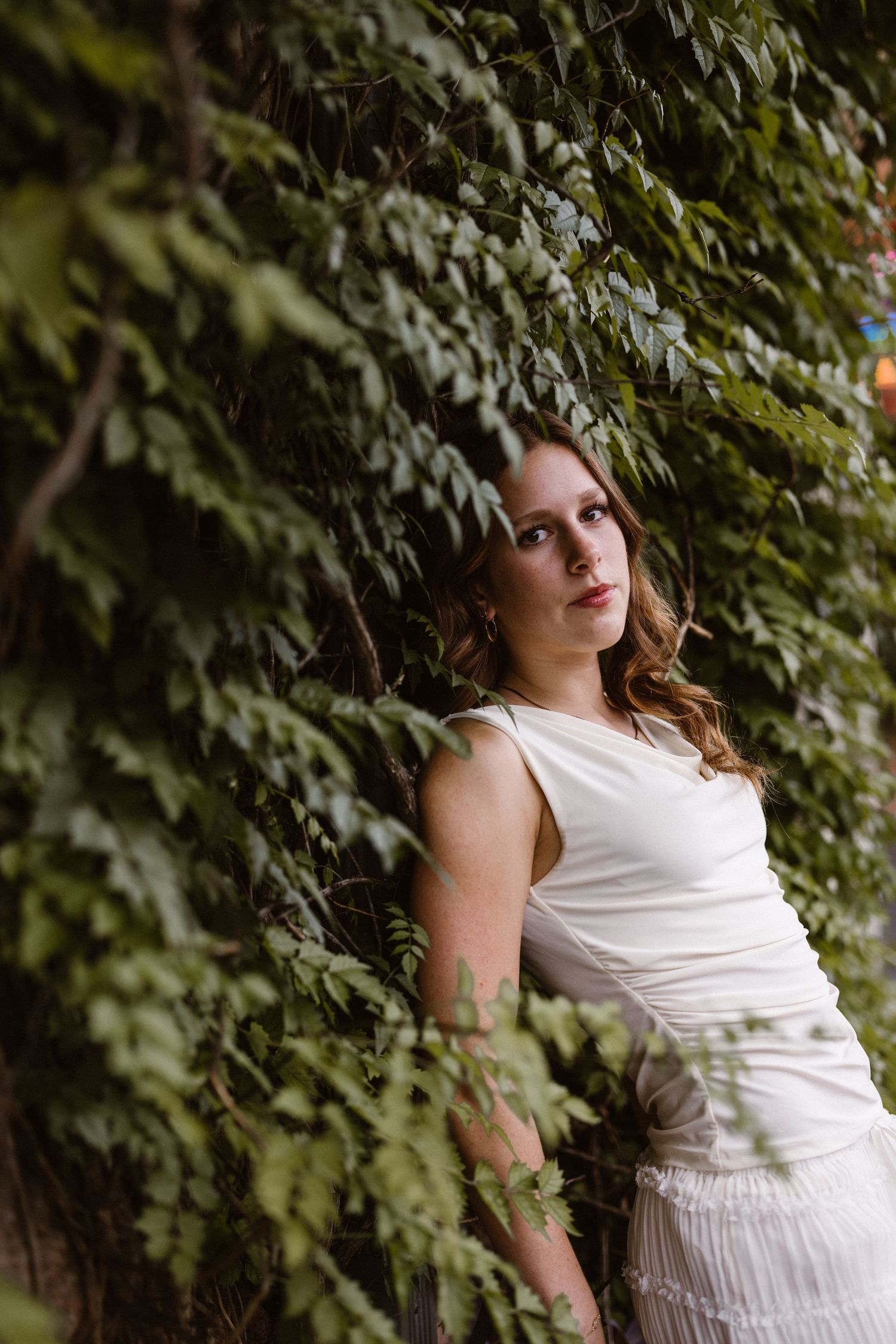 Young woman in a white sleeveless dress posing against a lush green ivy-covered wall outdoors.