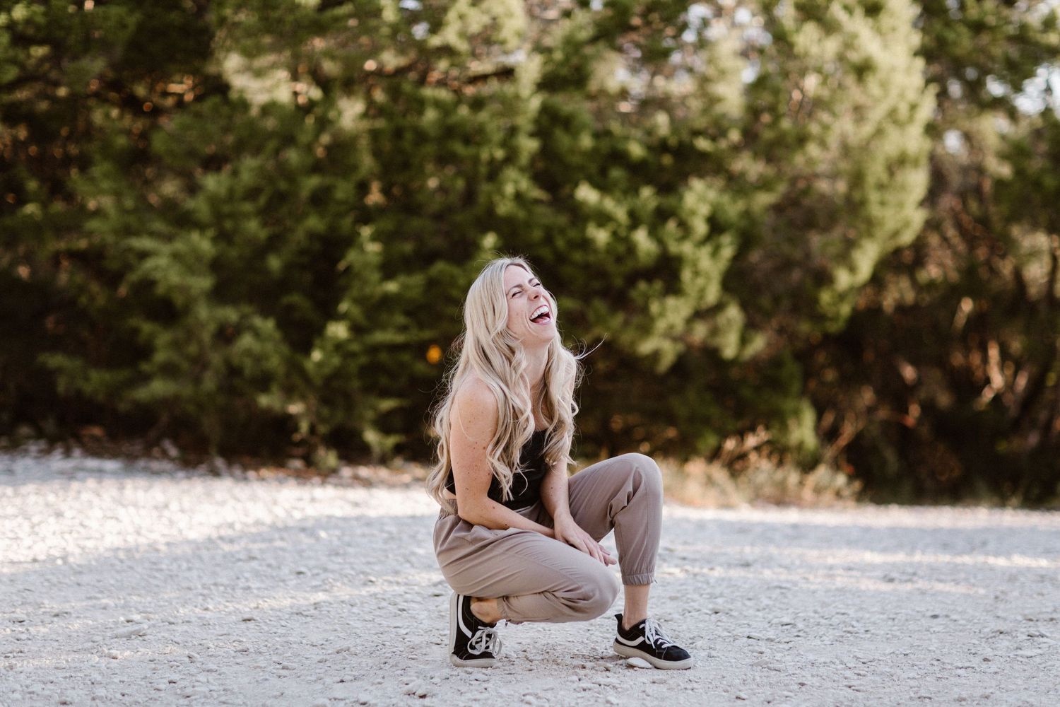 Blonde woman crouching on sandy ground laughing joyfully, wearing beige outfit and sneakers with green trees behind her.
