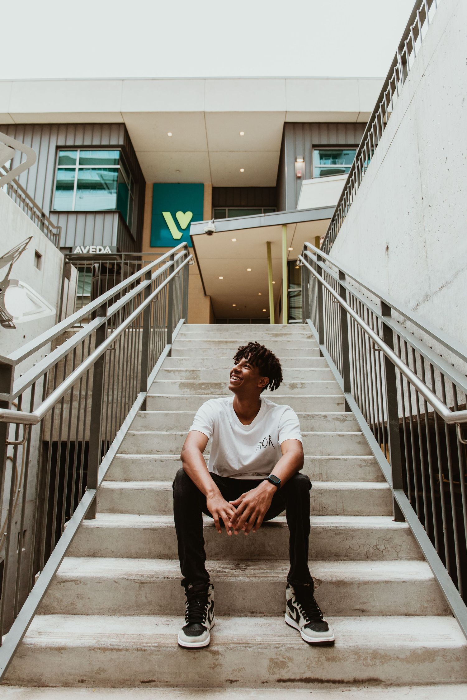 Young man in white tee and black jeans sitting on outdoor concrete stairs near an Aveda store.