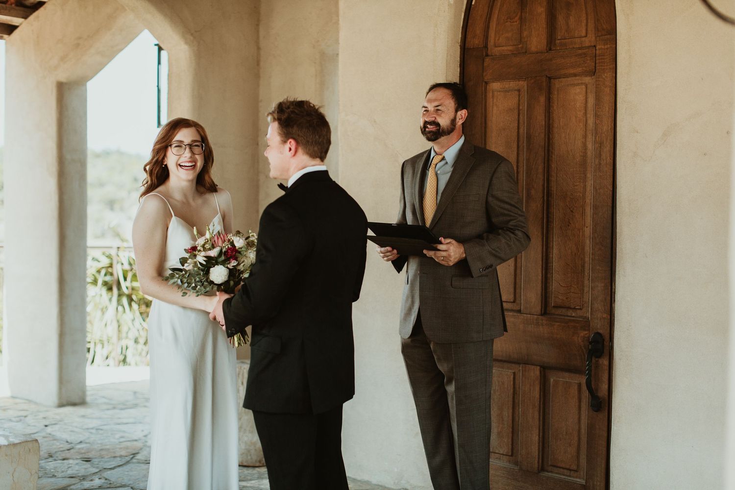 Bride and groom exchange vows with smiling officiant at rustic outdoor wedding ceremony near wooden door.