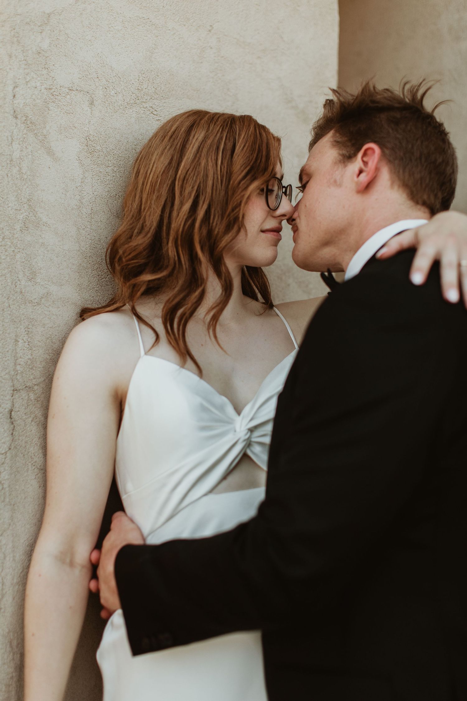 A bride in a white gown and groom in a black suit share an intimate moment, nearly kissing against a neutral wall.