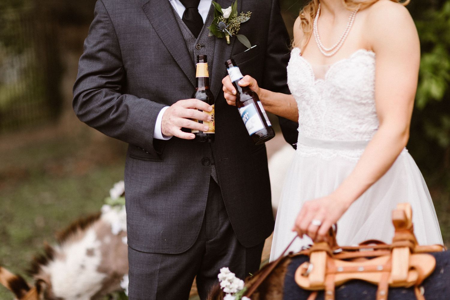A bride and groom hold beer bottles outdoors near a donkey at their wedding.