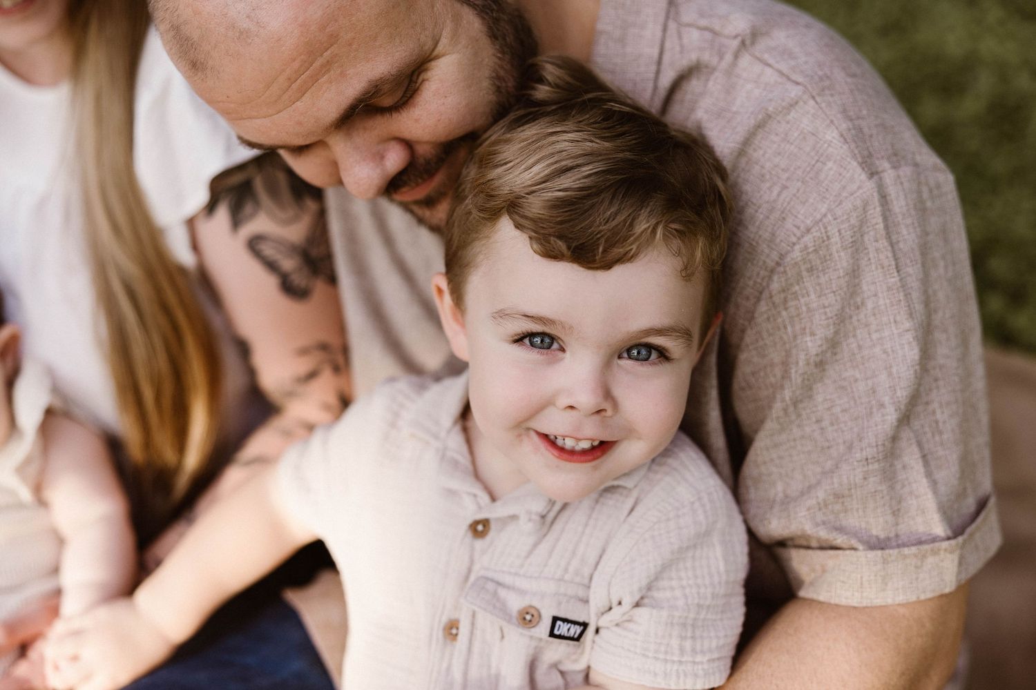 Smiling young boy with bright blue eyes being hugged by his father during an outdoor family session.