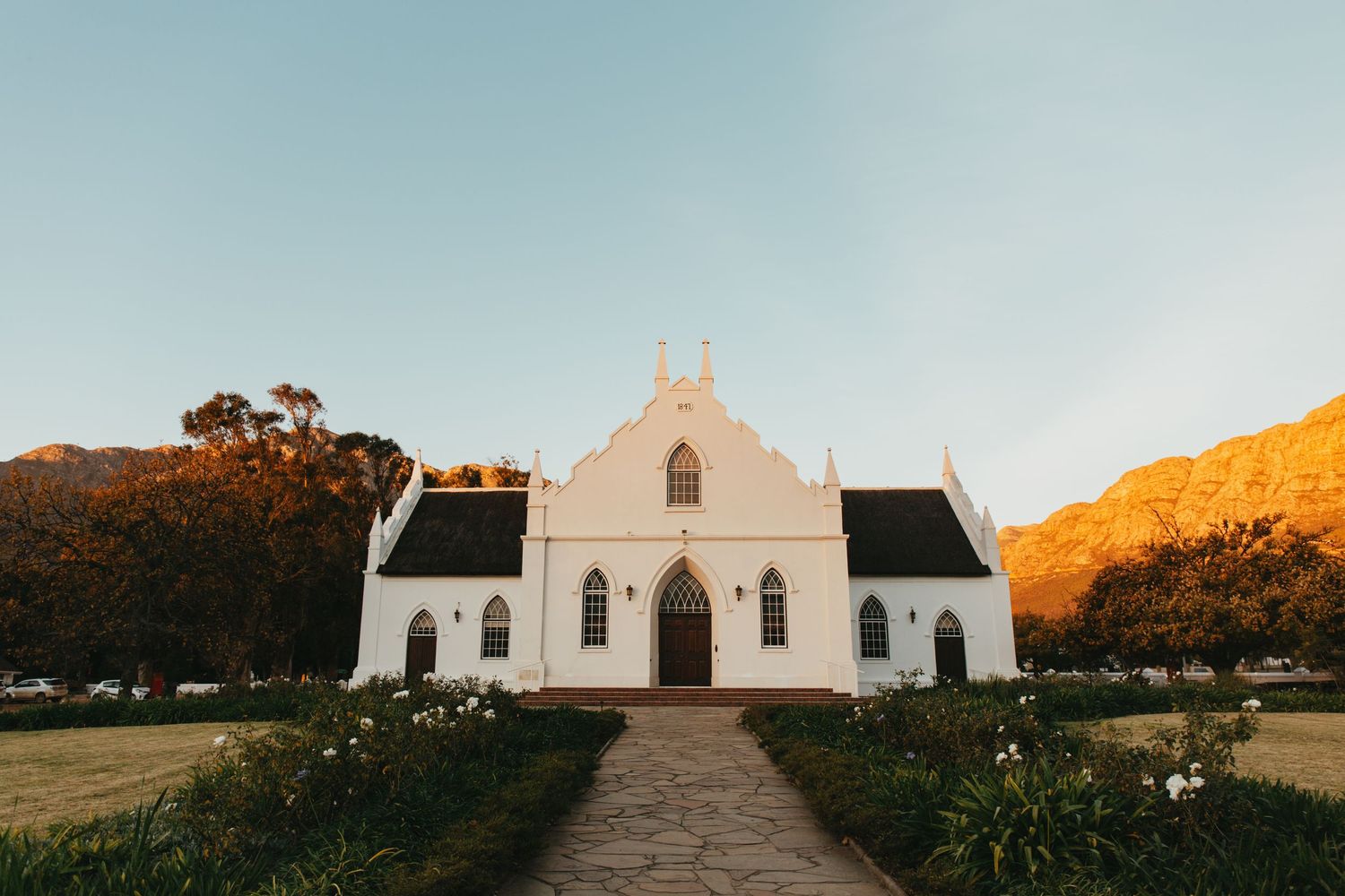 White Cape Dutch church with gabled facade, arched windows, and mountain backdrop at golden hour.