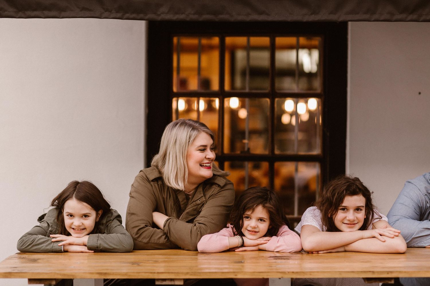 Mother and three children leaning on a wooden table smiling together outside a rustic building.