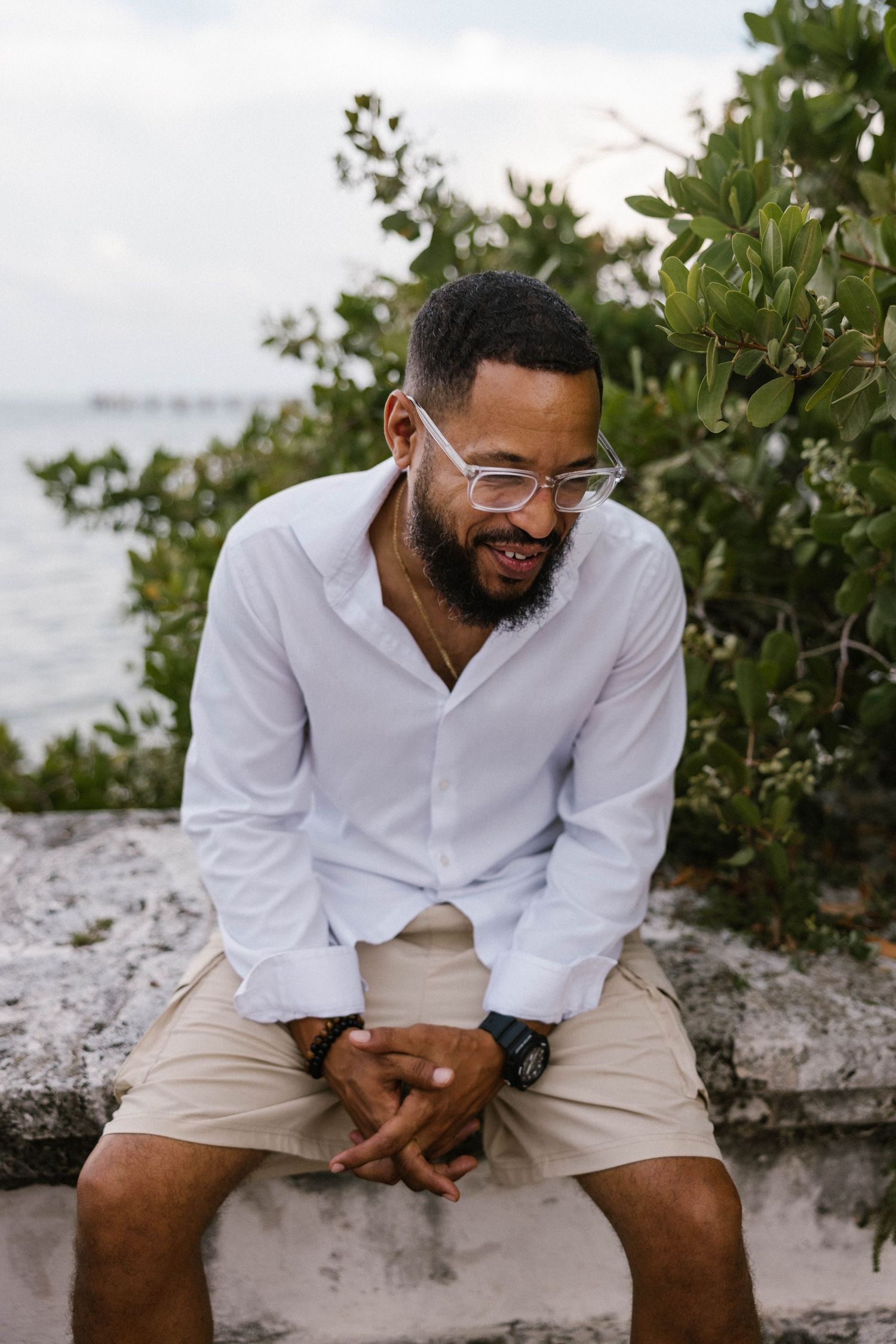 Smiling bearded man in white shirt and khaki shorts sits on a stone wall near the ocean.
