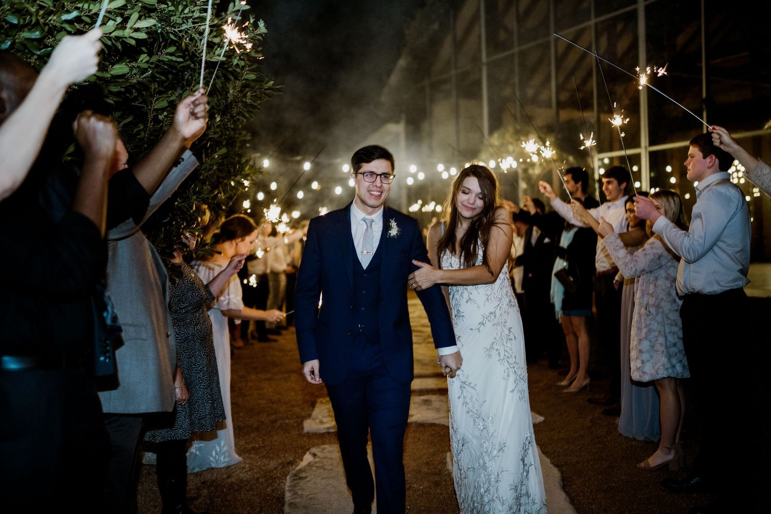 Newlywed couple walking through joyful sparkler exit tunnel surrounded by cheering guests at night wedding.