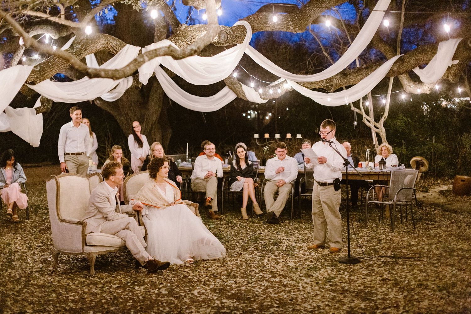A speaker addresses guests at a beautifully lit outdoor wedding reception under draped fabric lights.