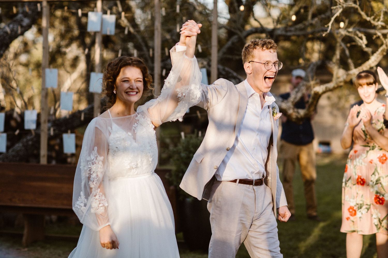 Laughing bride and groom holding hands raised in celebration at outdoor wedding with guests watching.