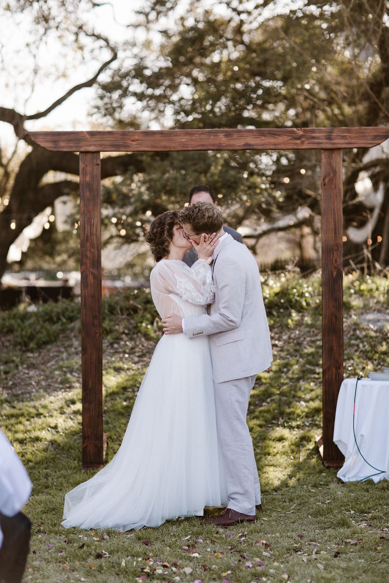 Bride and groom share first kiss under wooden arch at romantic outdoor wedding ceremony at golden hour.