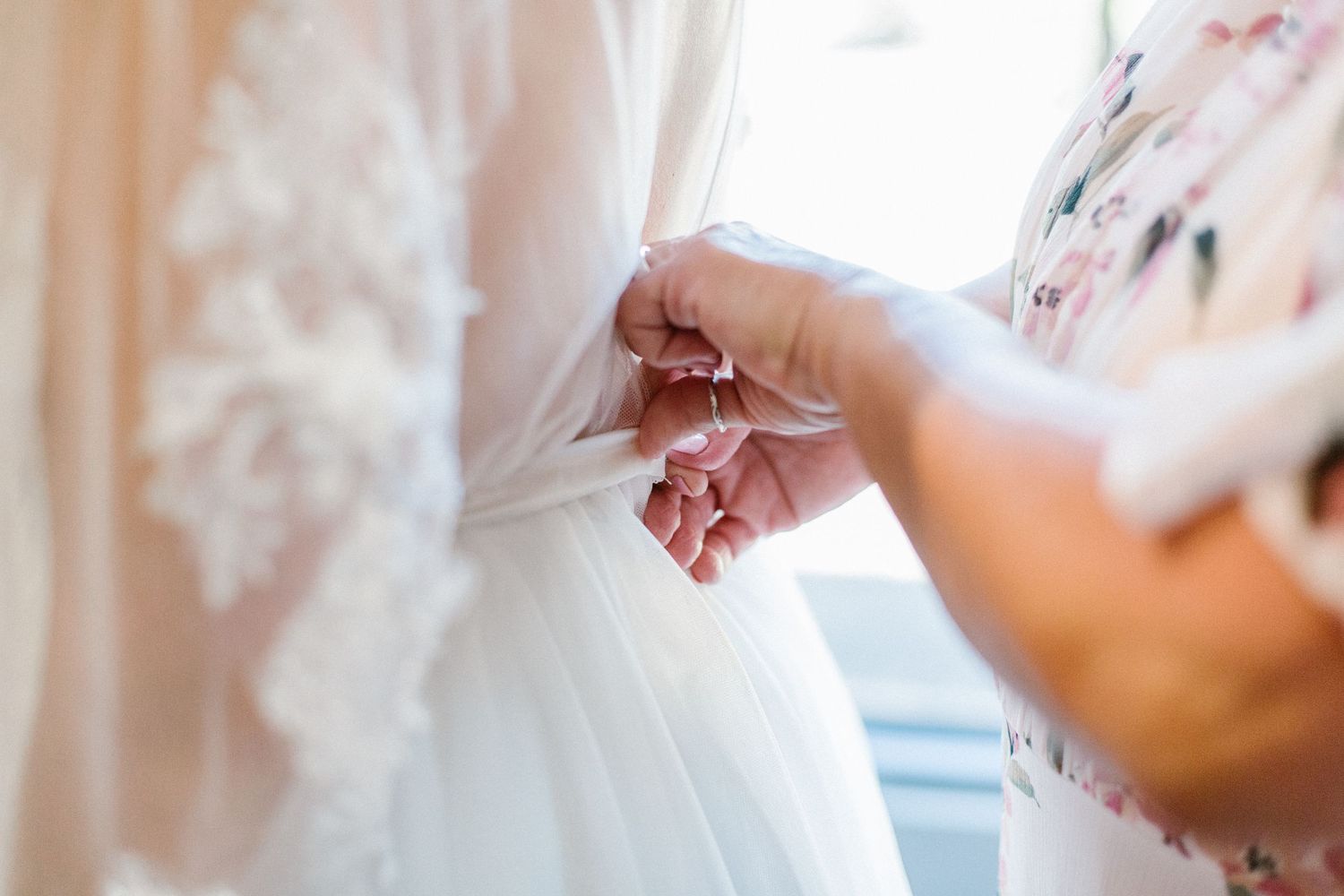 Close-up of hands fastening delicate buttons on the back of a lace wedding gown.