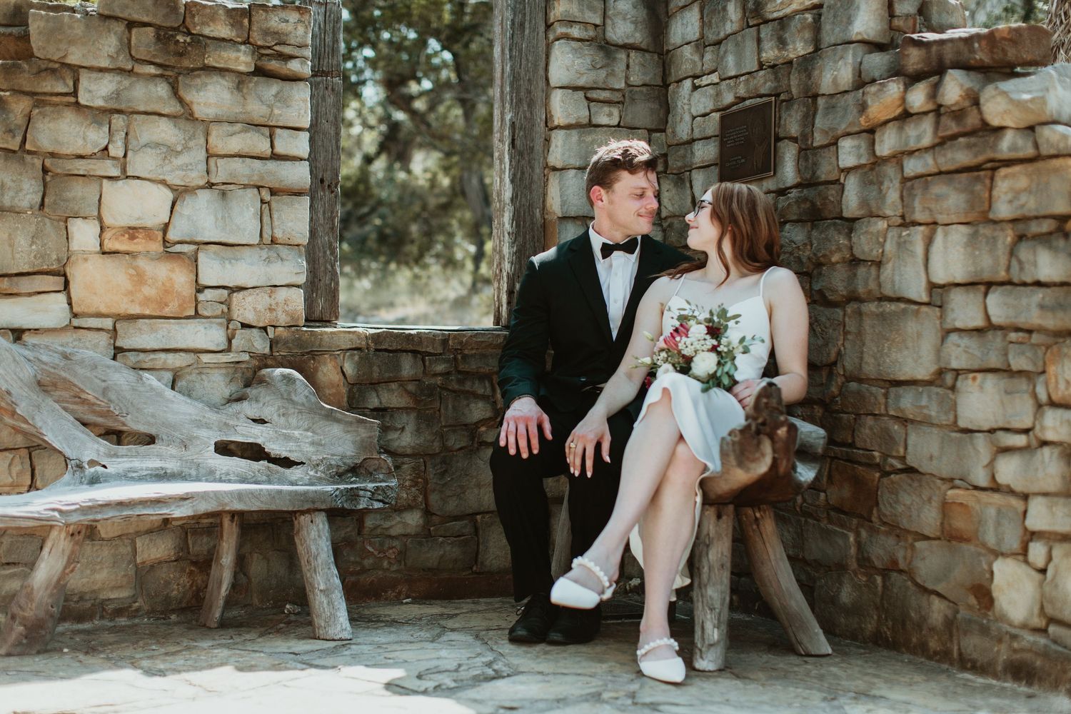 Bride and groom sitting together on rustic wooden bench against stone wall, bride holding bouquet.
