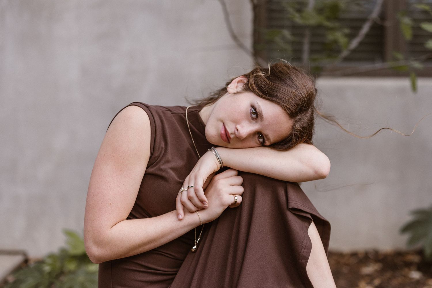Young woman in a brown sleeveless dress resting her head on her arms in a relaxed outdoor pose.