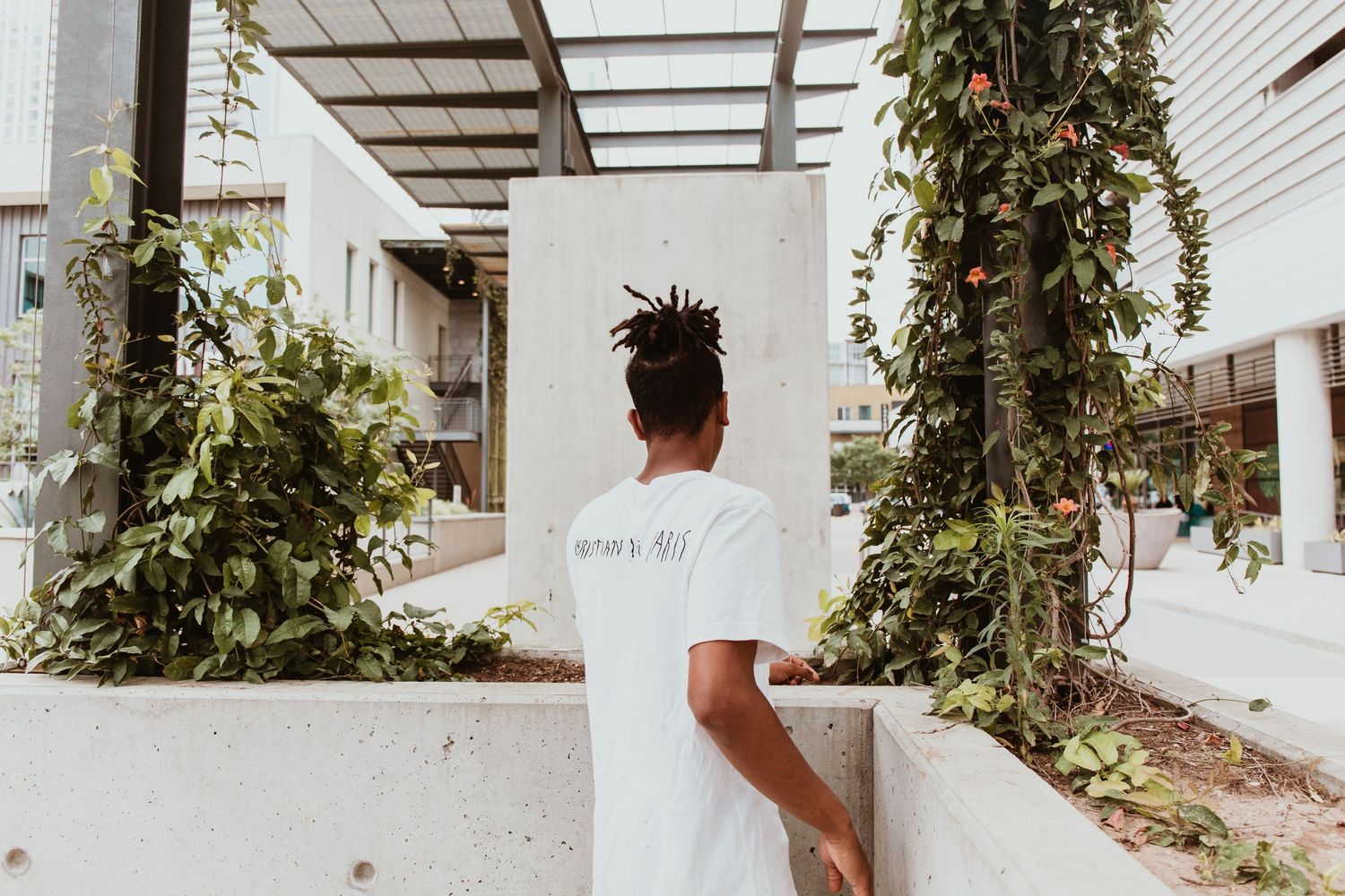 Young Black man with dreadlocks in white t-shirt stands near urban garden with lush green plants and modern building.