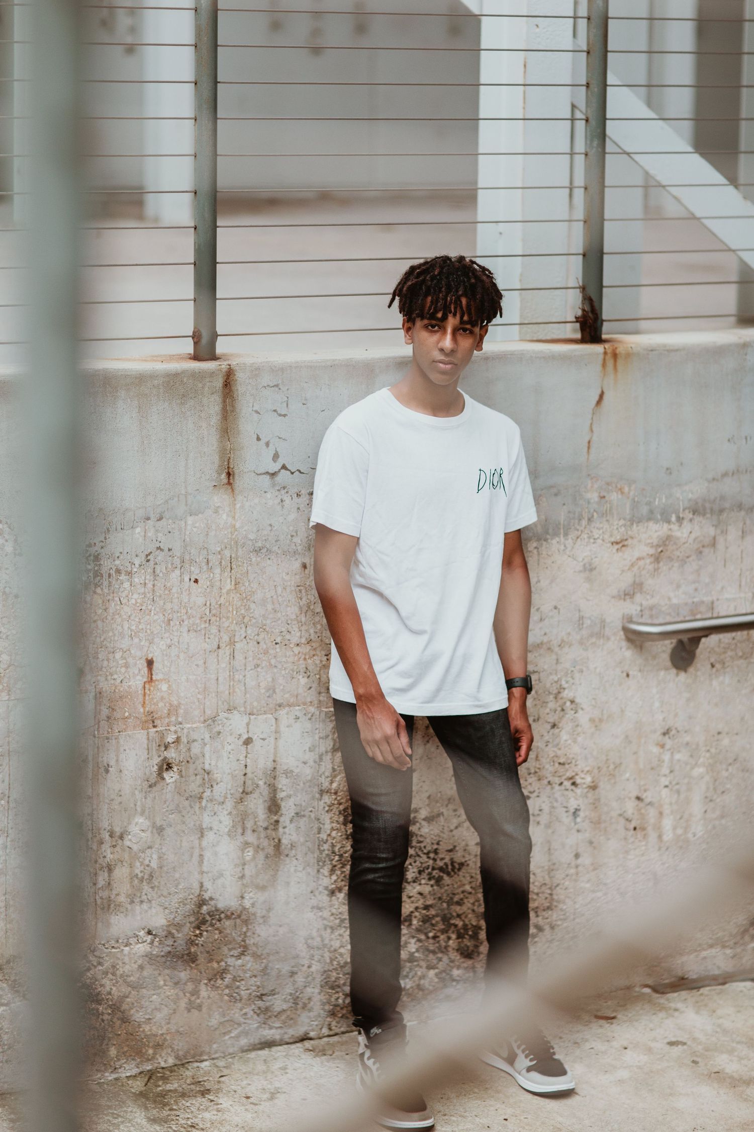Young man with dreadlocks wearing white t-shirt and gray jeans standing against weathered concrete wall outdoors.