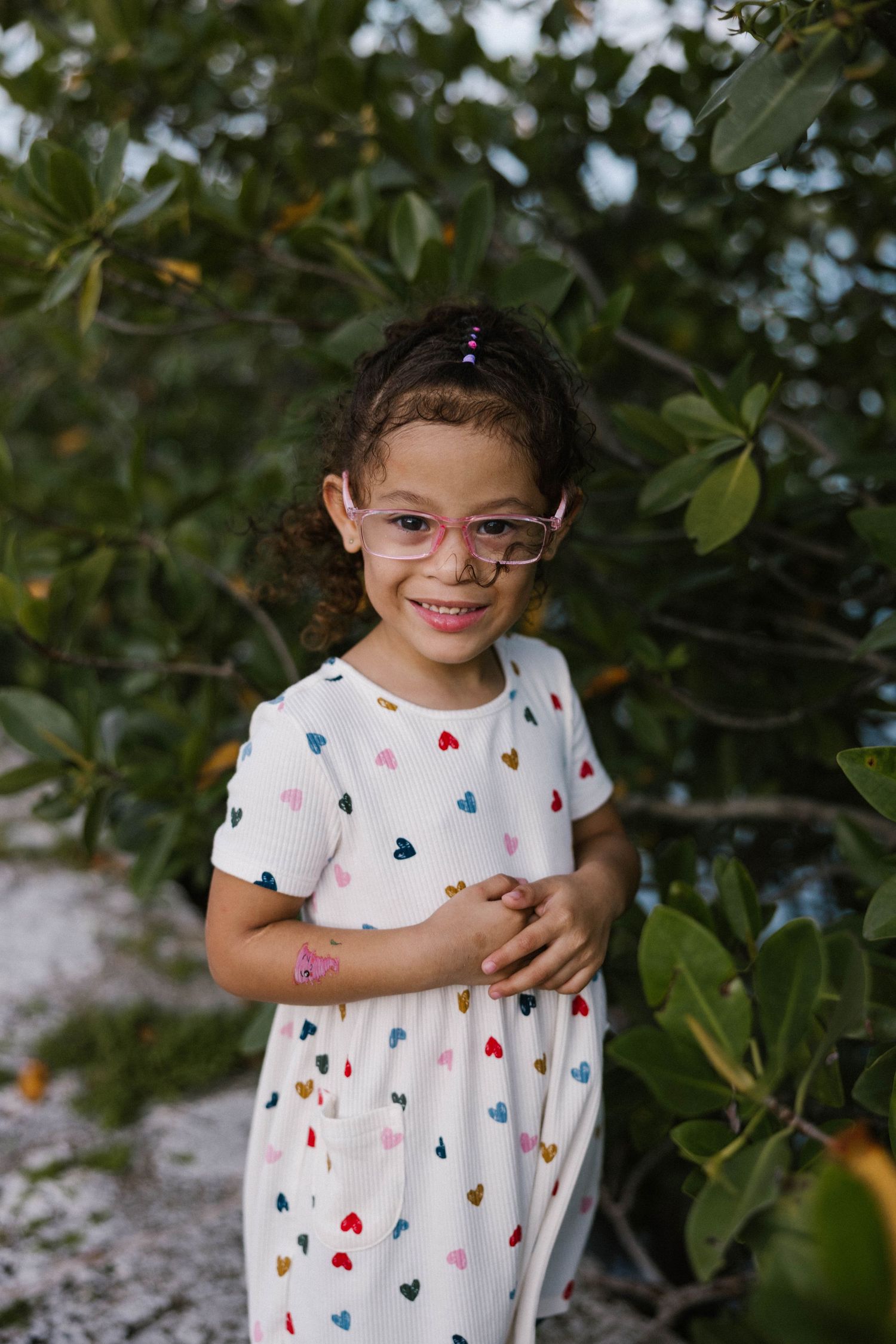 Young girl in heart-print dress and pink glasses smiles while standing near green foliage.