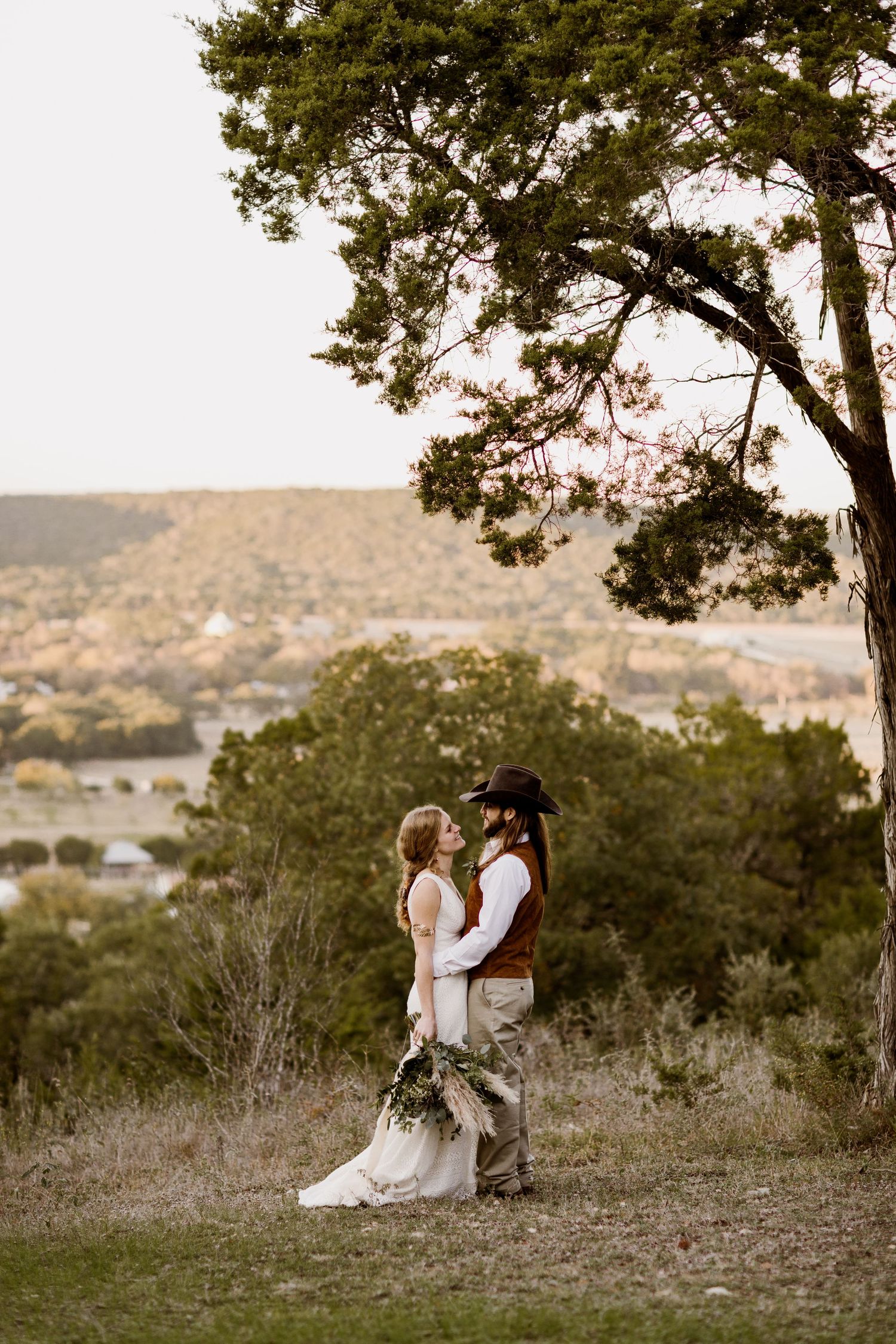 Bride and groom stand facing each other on hilltop at sunset with scenic Texas Hill Country backdrop.