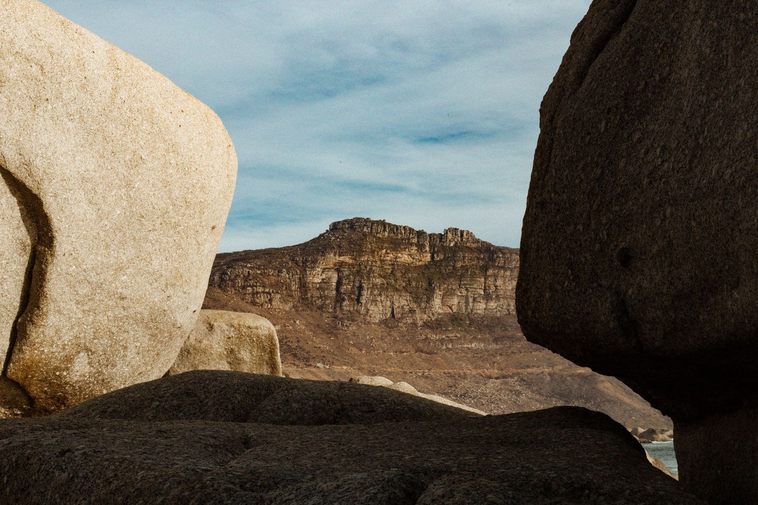Large granite boulders frame a dramatic mesa cliff face under a partly cloudy sky in a rugged coastal landscape.