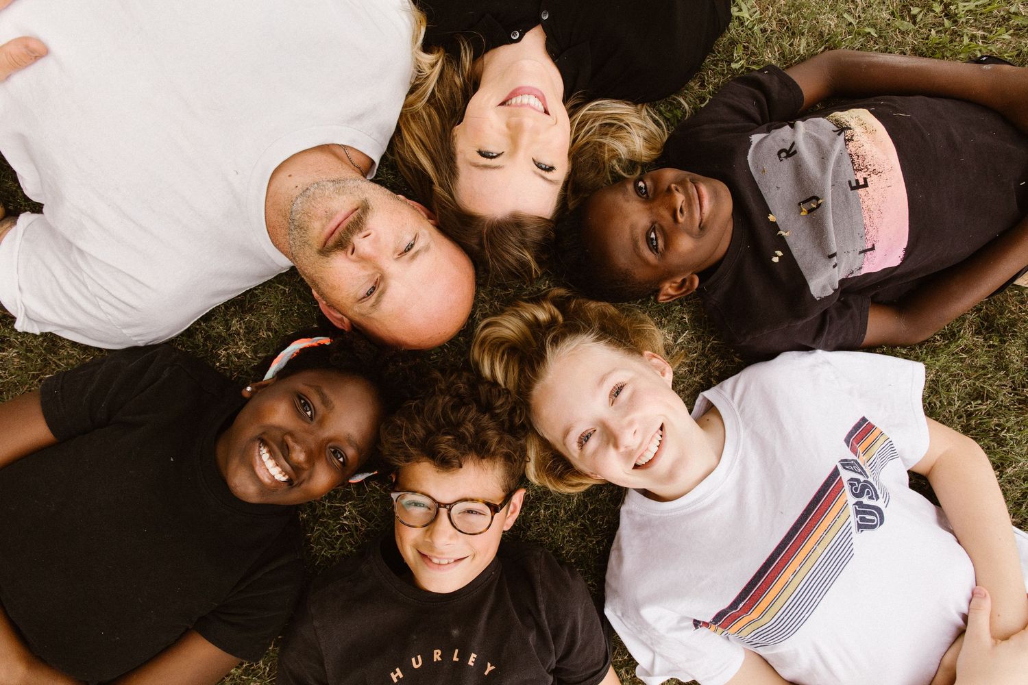Six diverse friends lying in a circle on grass, laughing and smiling together.