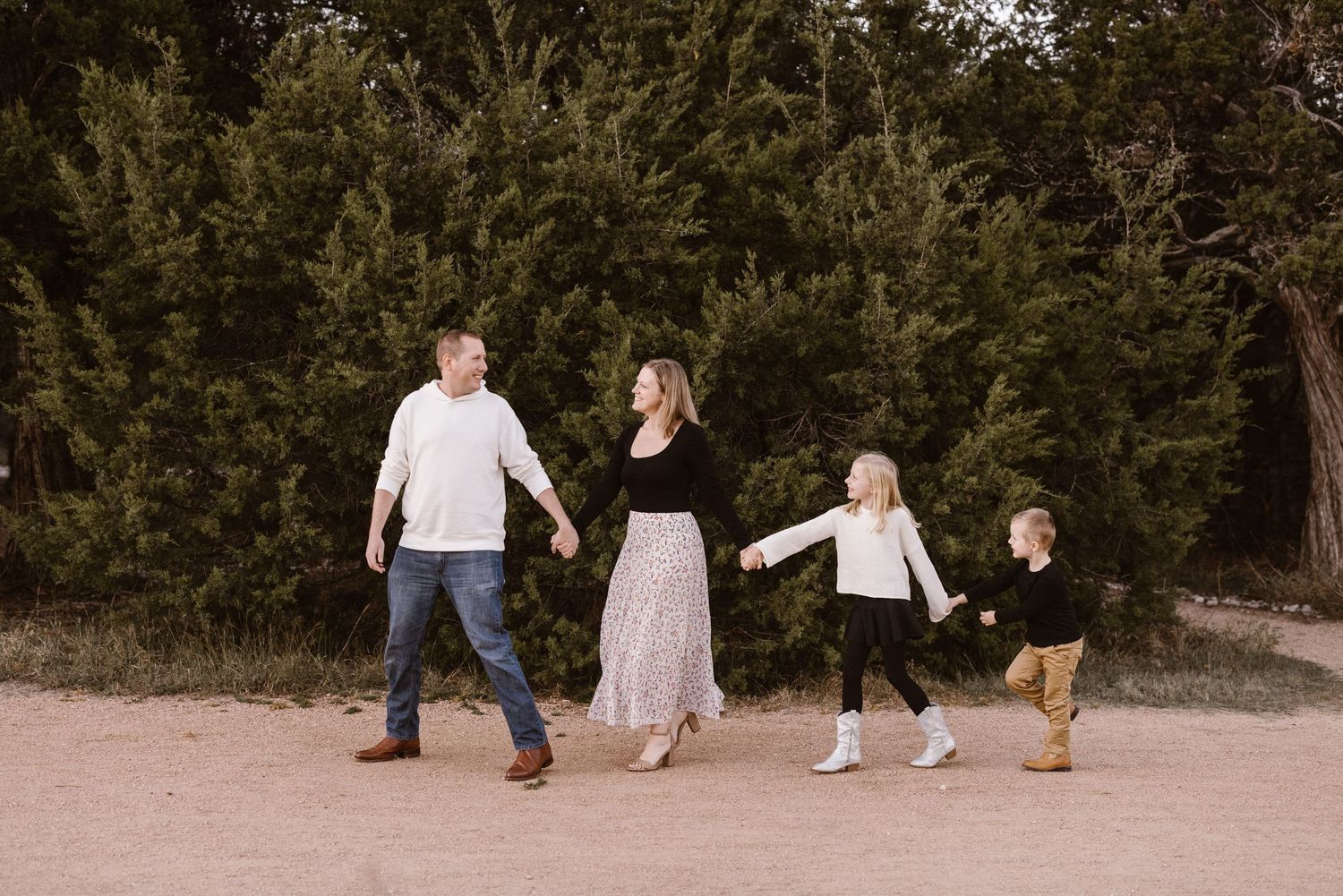 Family of four walks hand in hand along a dirt path surrounded by evergreen trees.