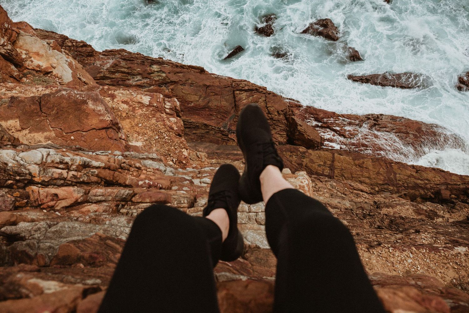 Person sitting on rocky coastal cliff edge with legs dangling over crashing ocean waves below.