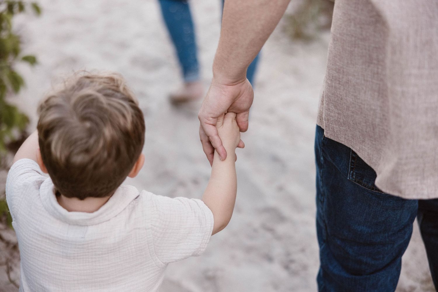 A young child holds an adult's hand while walking on a sandy beach path.