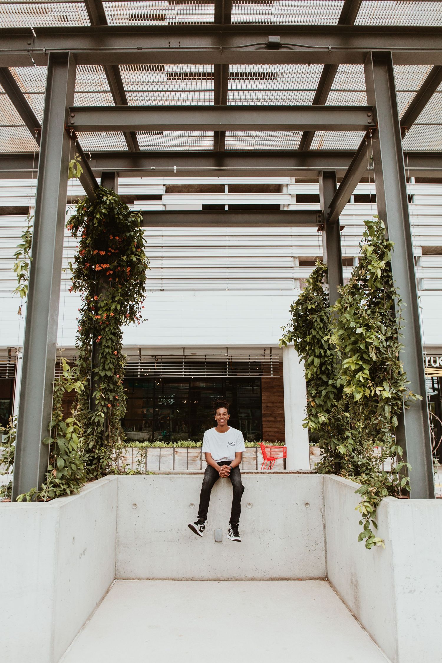 Young man sitting on white concrete ledge between vine-covered pillars under modern pergola.