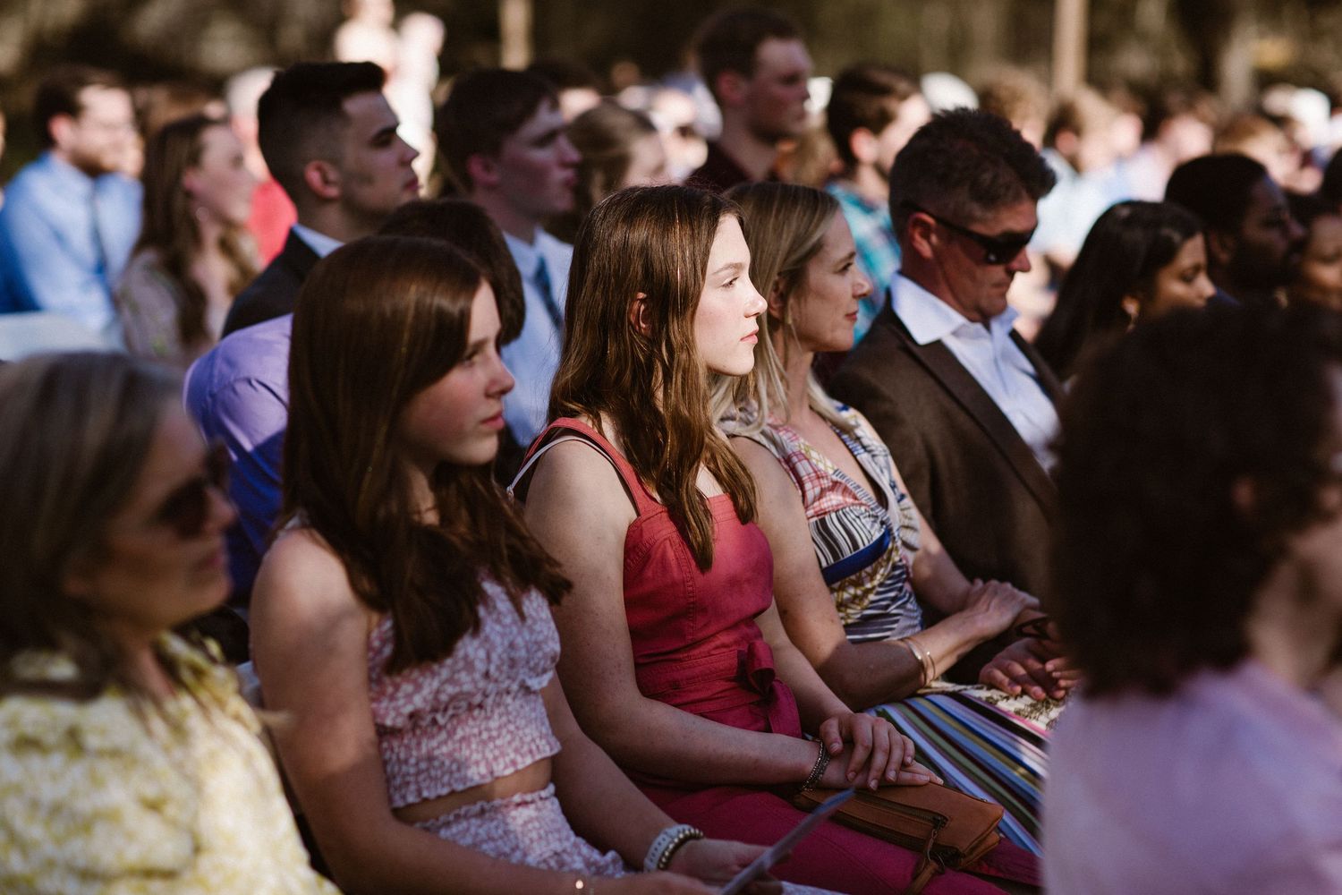 Elegantly dressed women seated in the front row attentively watching an outdoor wedding ceremony.