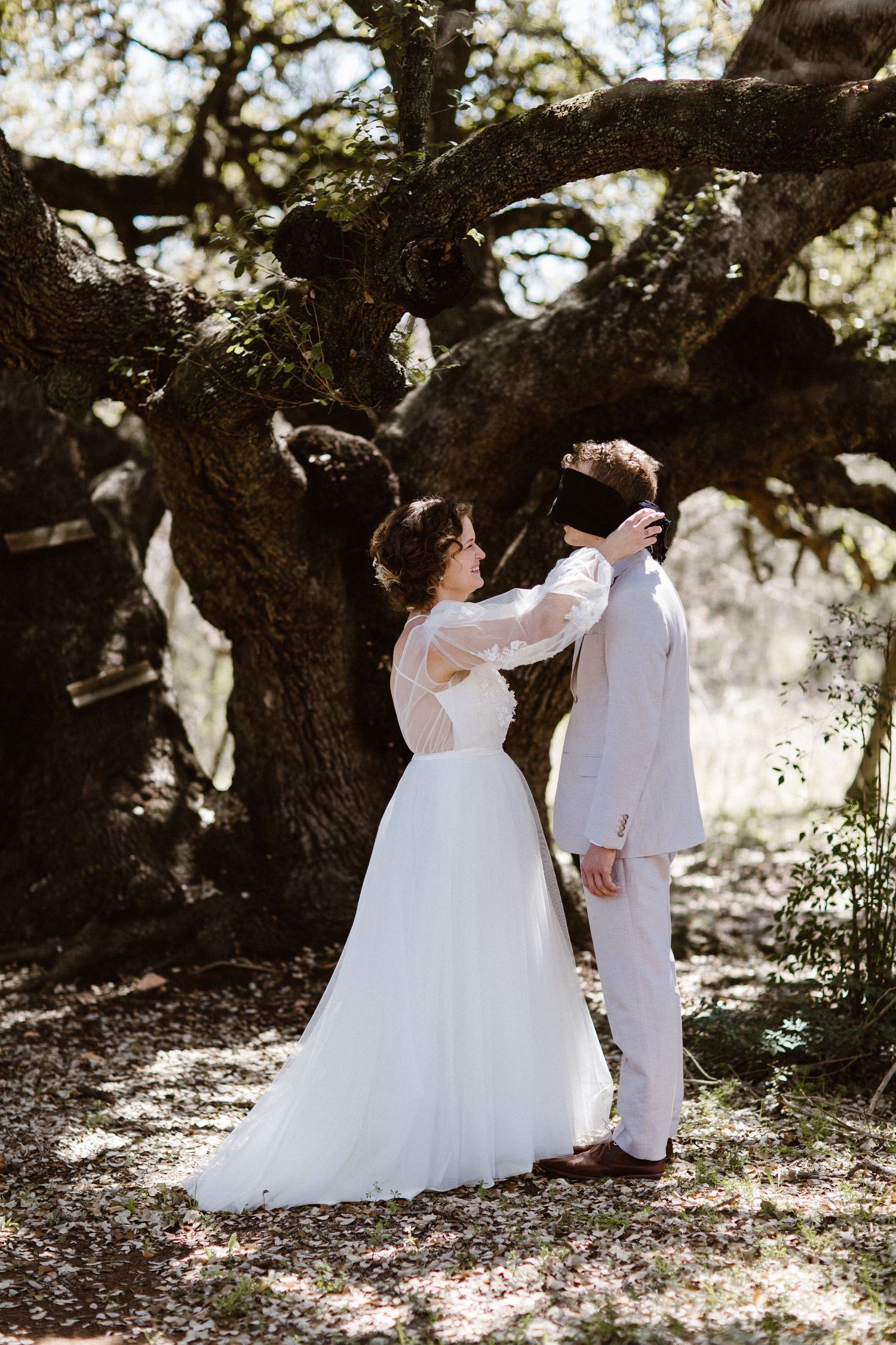 Bride removing blindfold from groom during an emotional first look under a majestic oak tree.