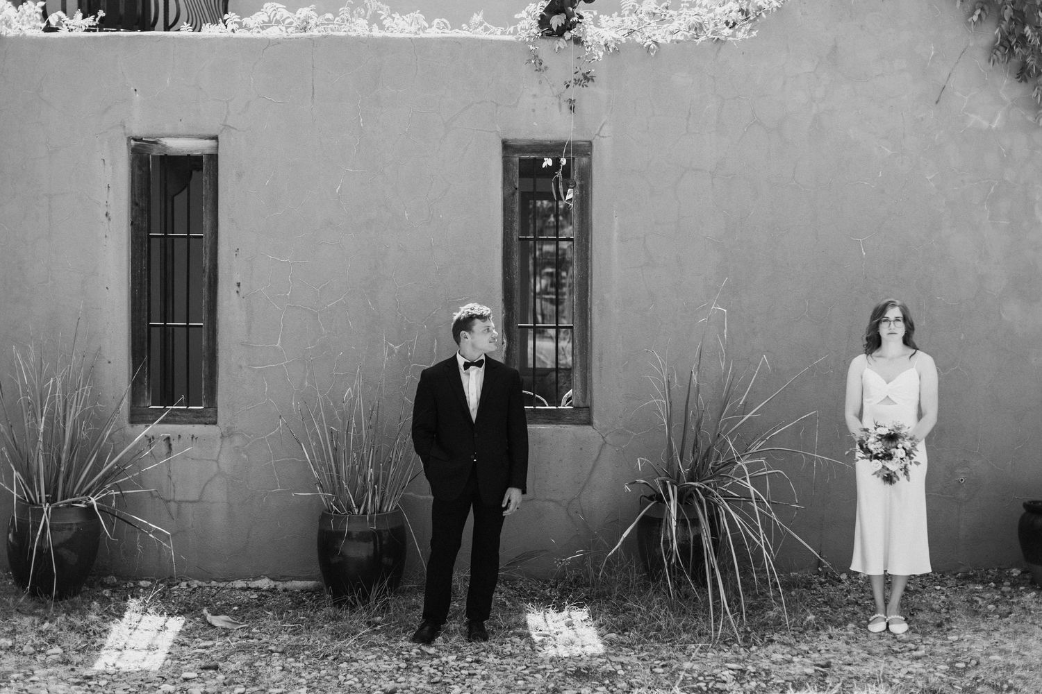 Black and white wedding photo of bride and groom standing apart against adobe wall outdoors.