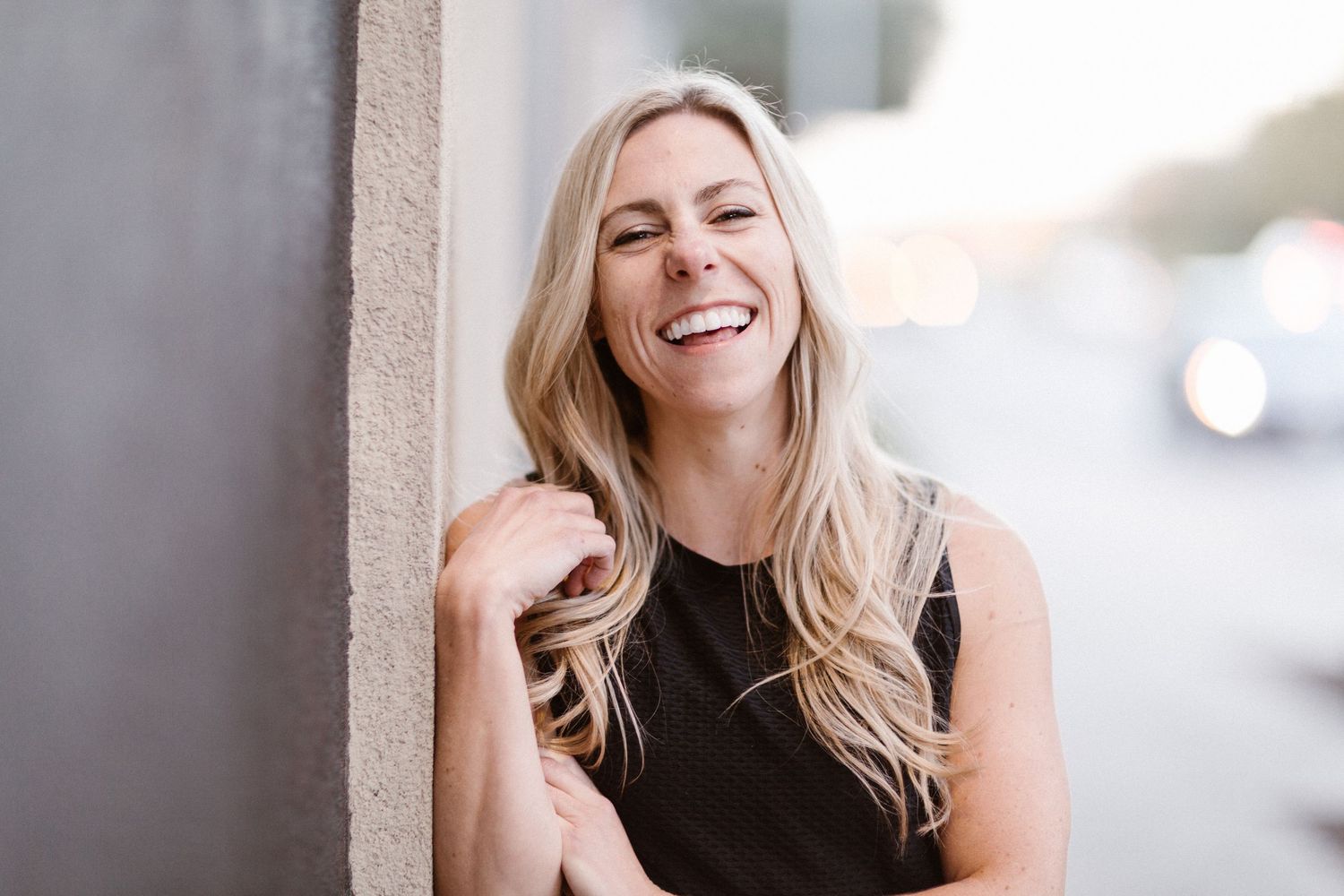 Smiling blonde woman leaning against a wall outdoors, wearing a black top, laughing joyfully in natural light.