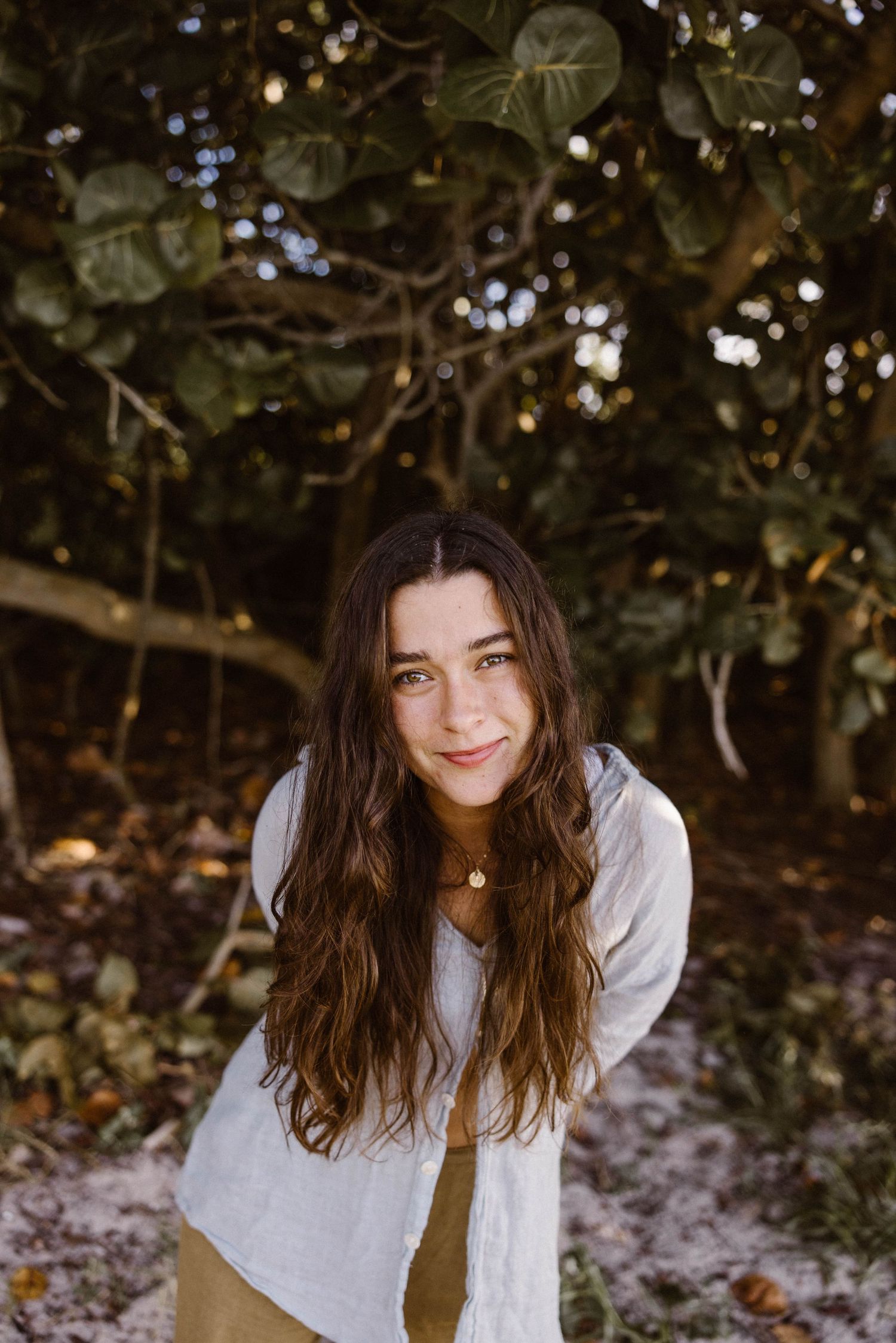 Smiling young woman with long wavy brown hair wearing a white blouse standing in front of lush green trees.