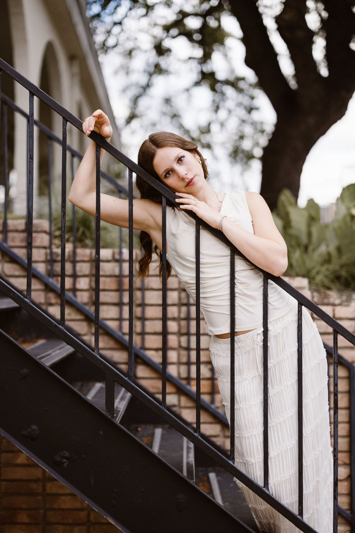 Young woman in a white outfit leaning on black metal stair railing beside a brick building outdoors.