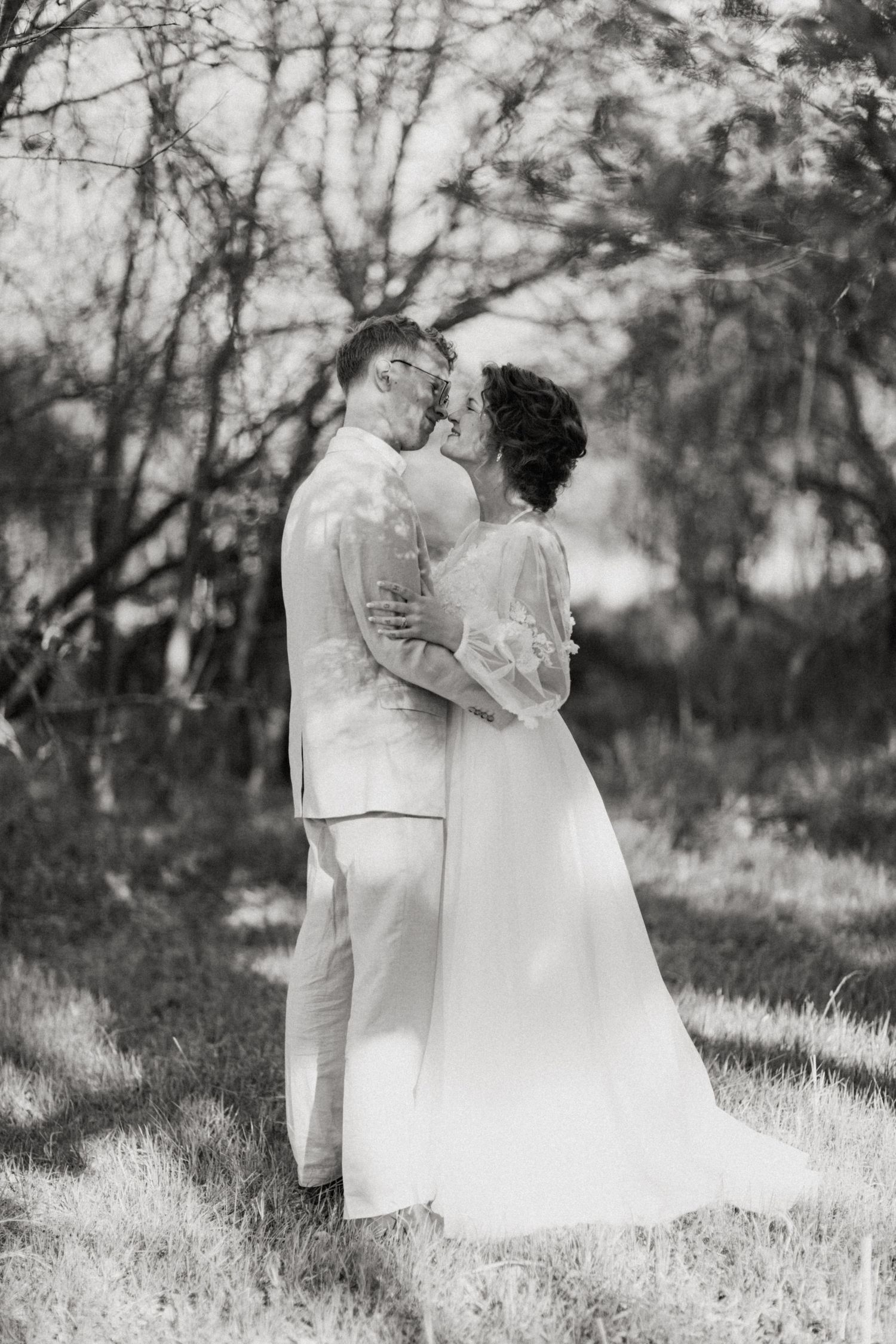 Black and white romantic photo of bride and groom kissing while embracing in outdoor wooded setting.