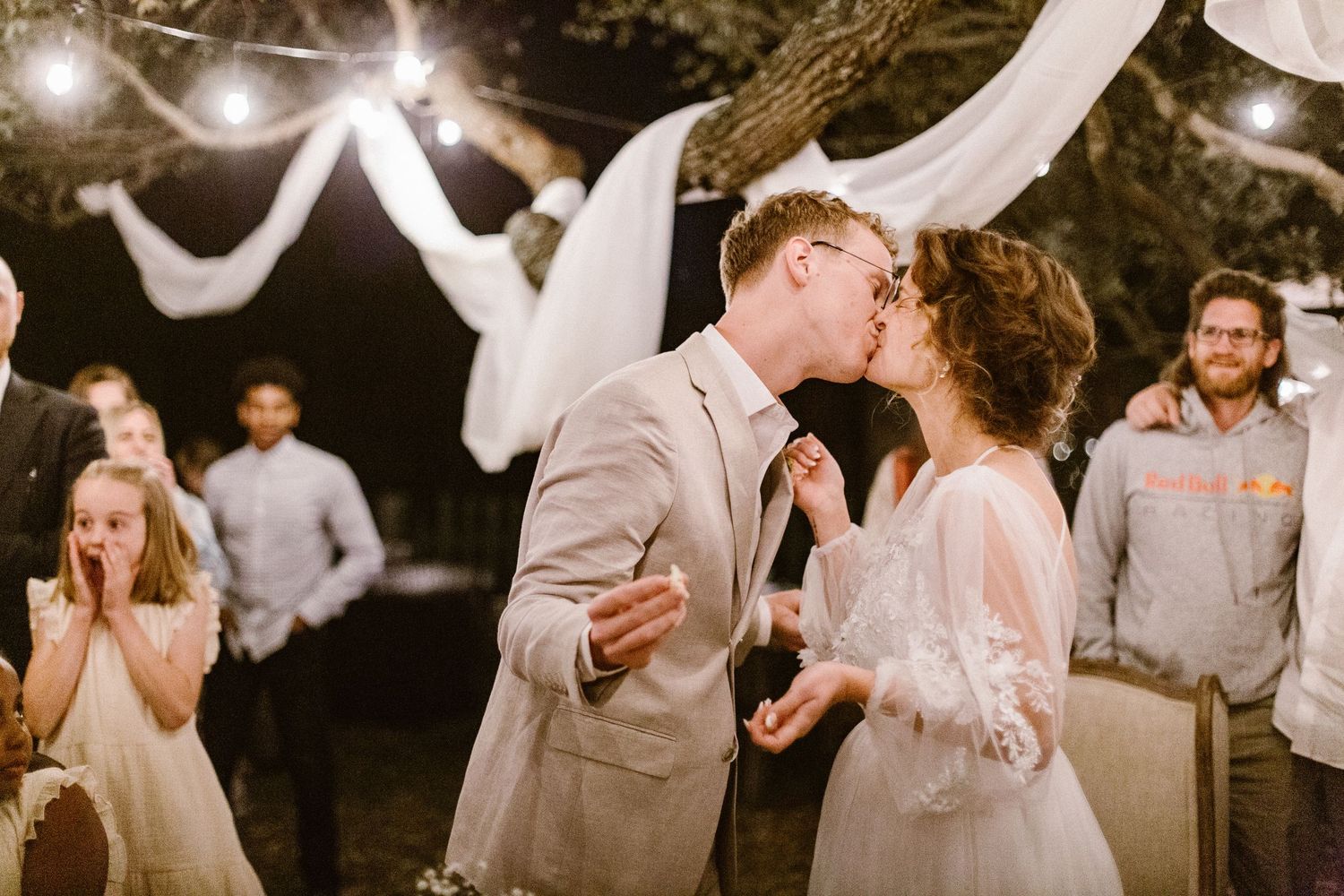 Newlyweds sharing first kiss at outdoor wedding reception under warm string lights.