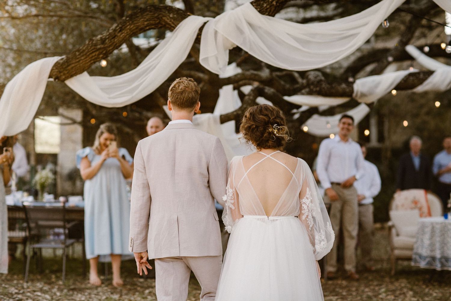 Bride and groom walking hand in hand toward outdoor wedding reception decorated with white draping and string lights.