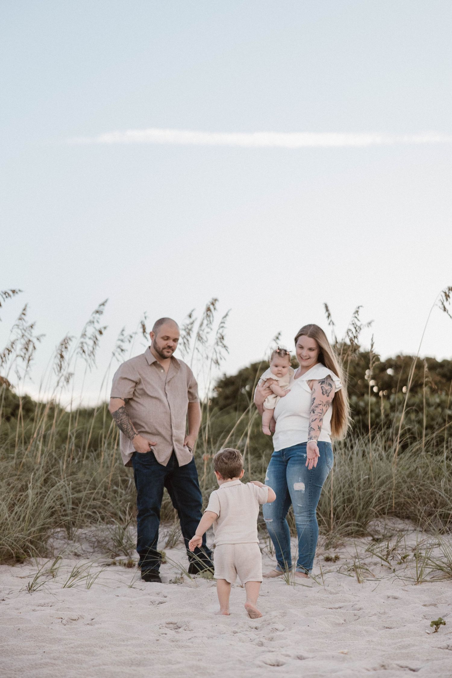 Family of four enjoying a beach day near sand dunes with sea oats, toddler running toward parents holding baby.