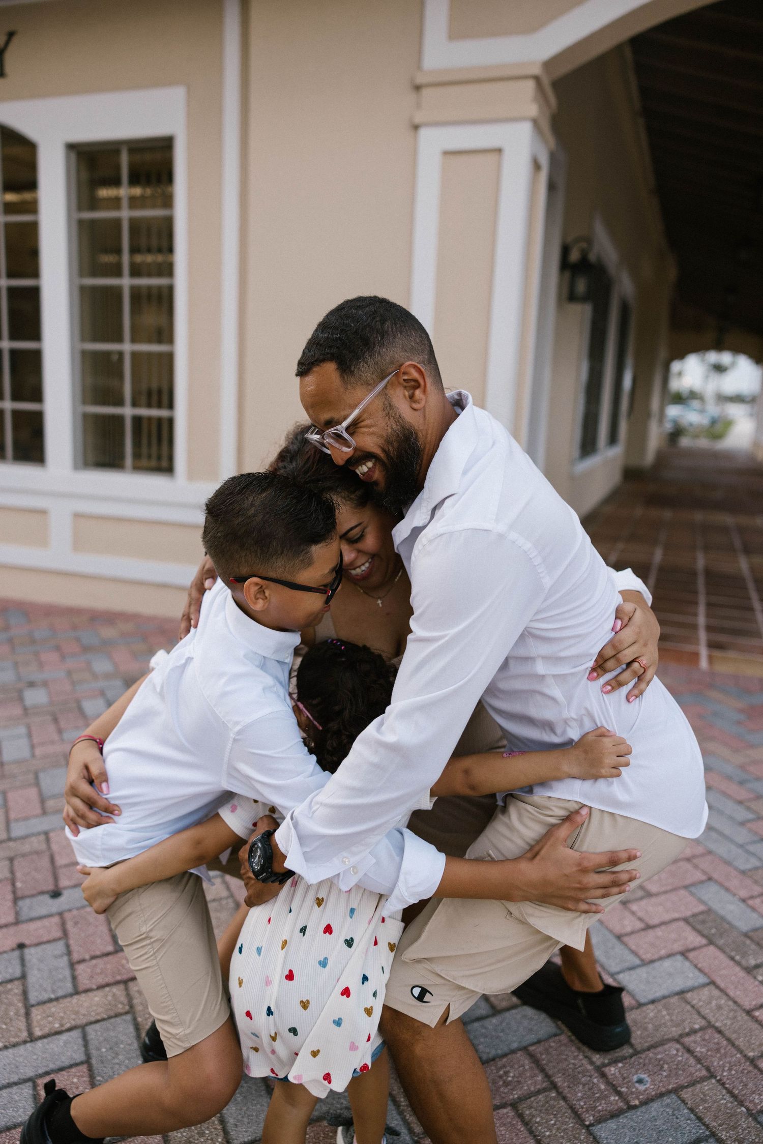 Family of four shares a warm group hug on a brick walkway outside a building.