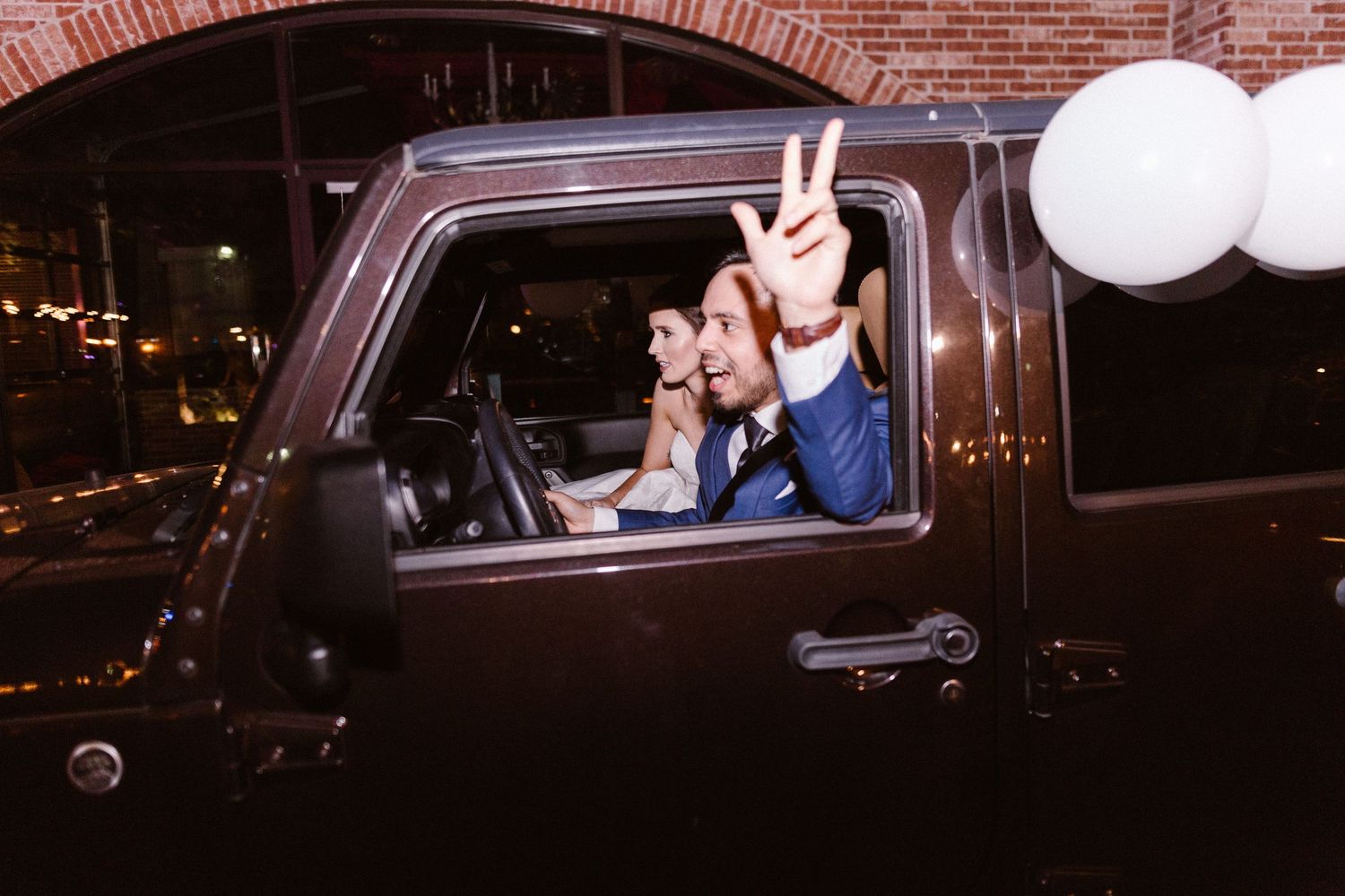 Groom flashing peace sign from dark truck window during festive wedding send-off.