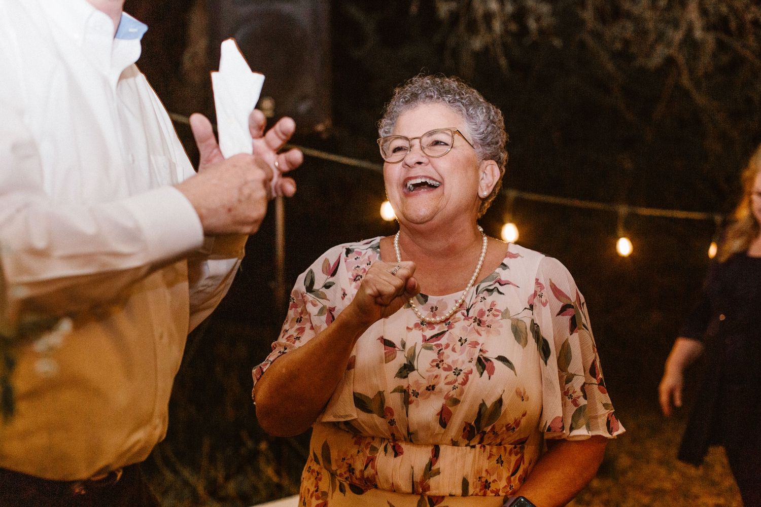 Older woman in floral dress laughing warmly while holding a candle at a party.