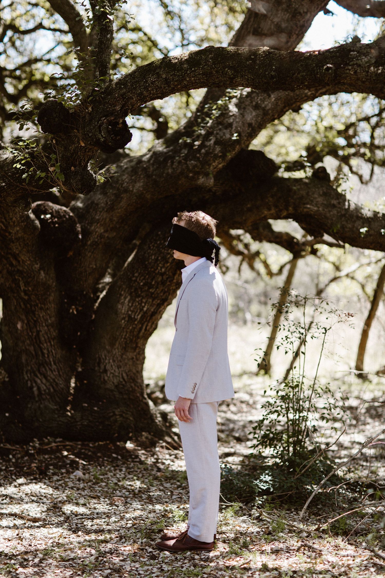 Blindfolded groom in a light grey suit standing beneath a large oak tree outdoors.