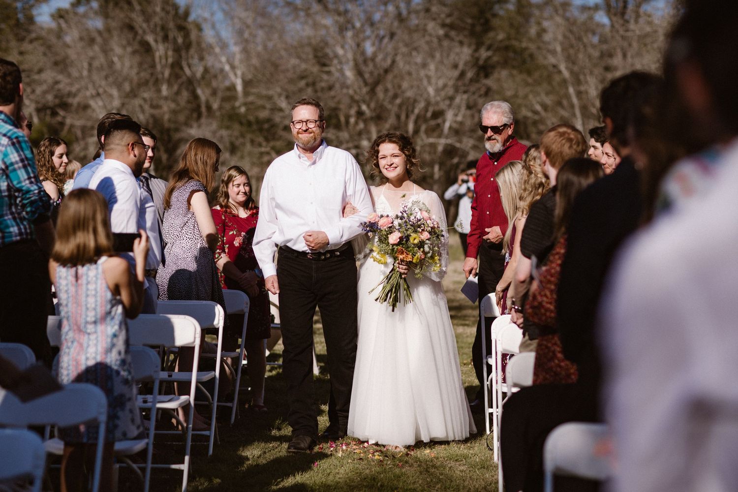 Bride in white gown holding a colorful bouquet walks down the aisle with her father during an outdoor wedding ceremony.