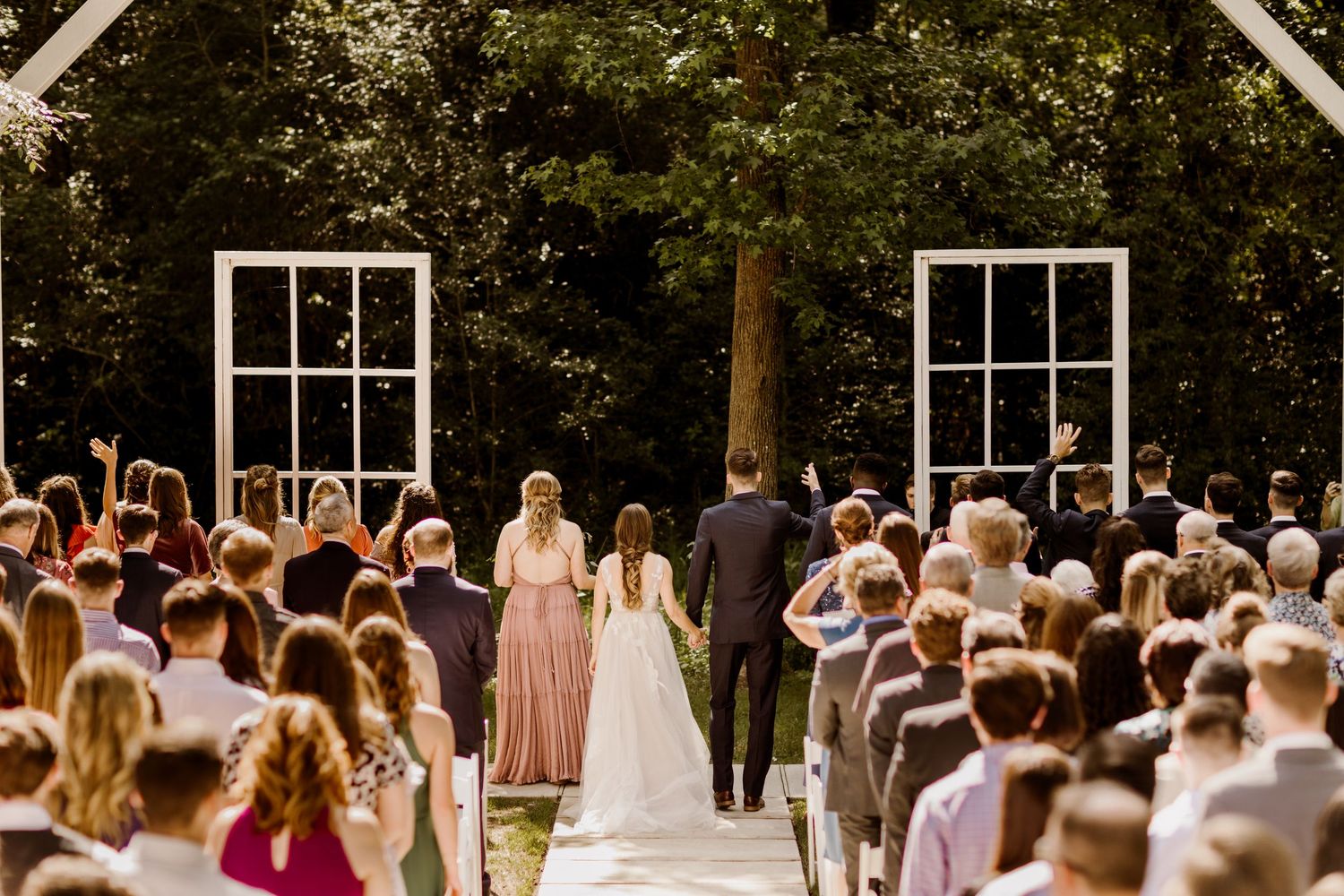 Outdoor wedding ceremony with guests standing as bride walks down aisle toward white window frame altar in lush garden.