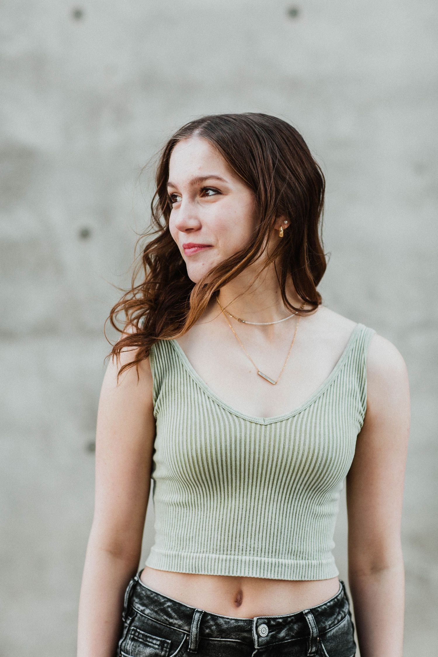 Young woman in a sage green ribbed crop top and dark jeans posing against a concrete wall outdoors.