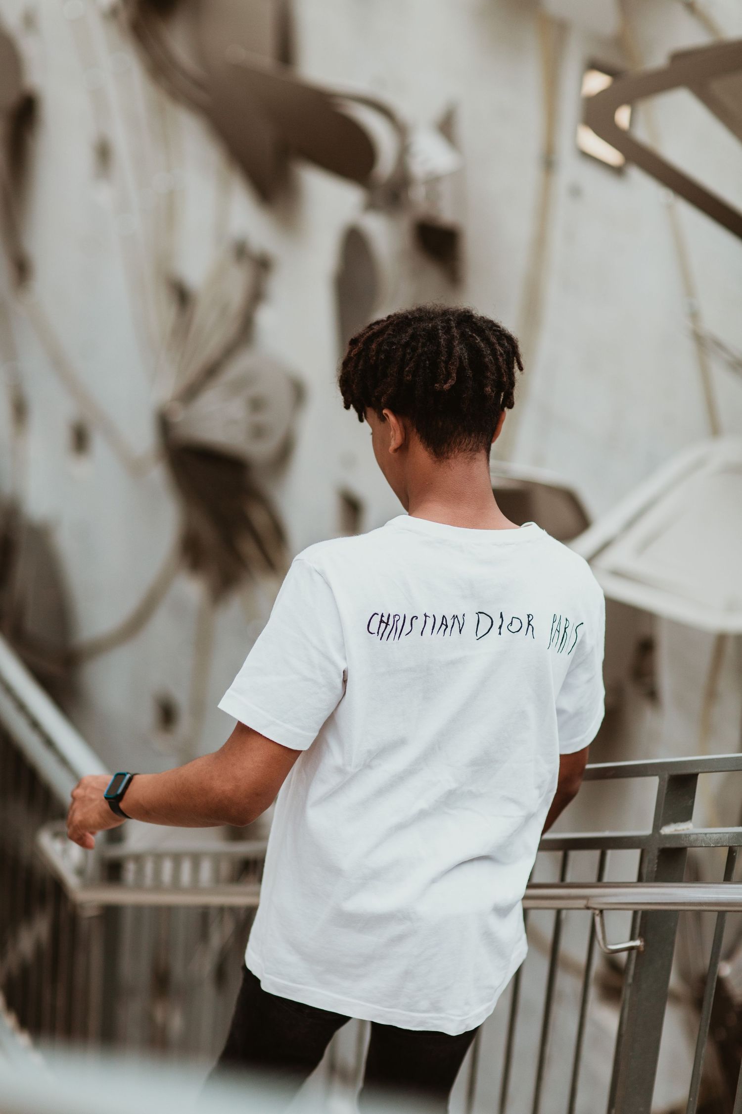 Young man in white Christian Dior t-shirt viewed from behind near metal railing.