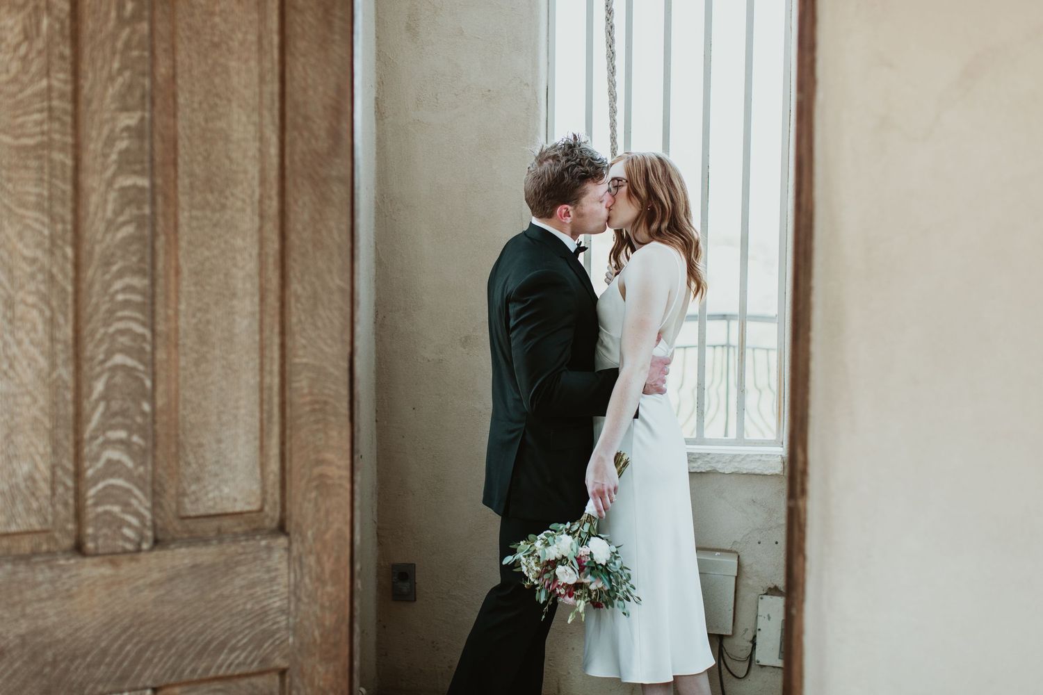A bride and groom share a romantic kiss near a window in a rustic venue, bride holding a bouquet.
