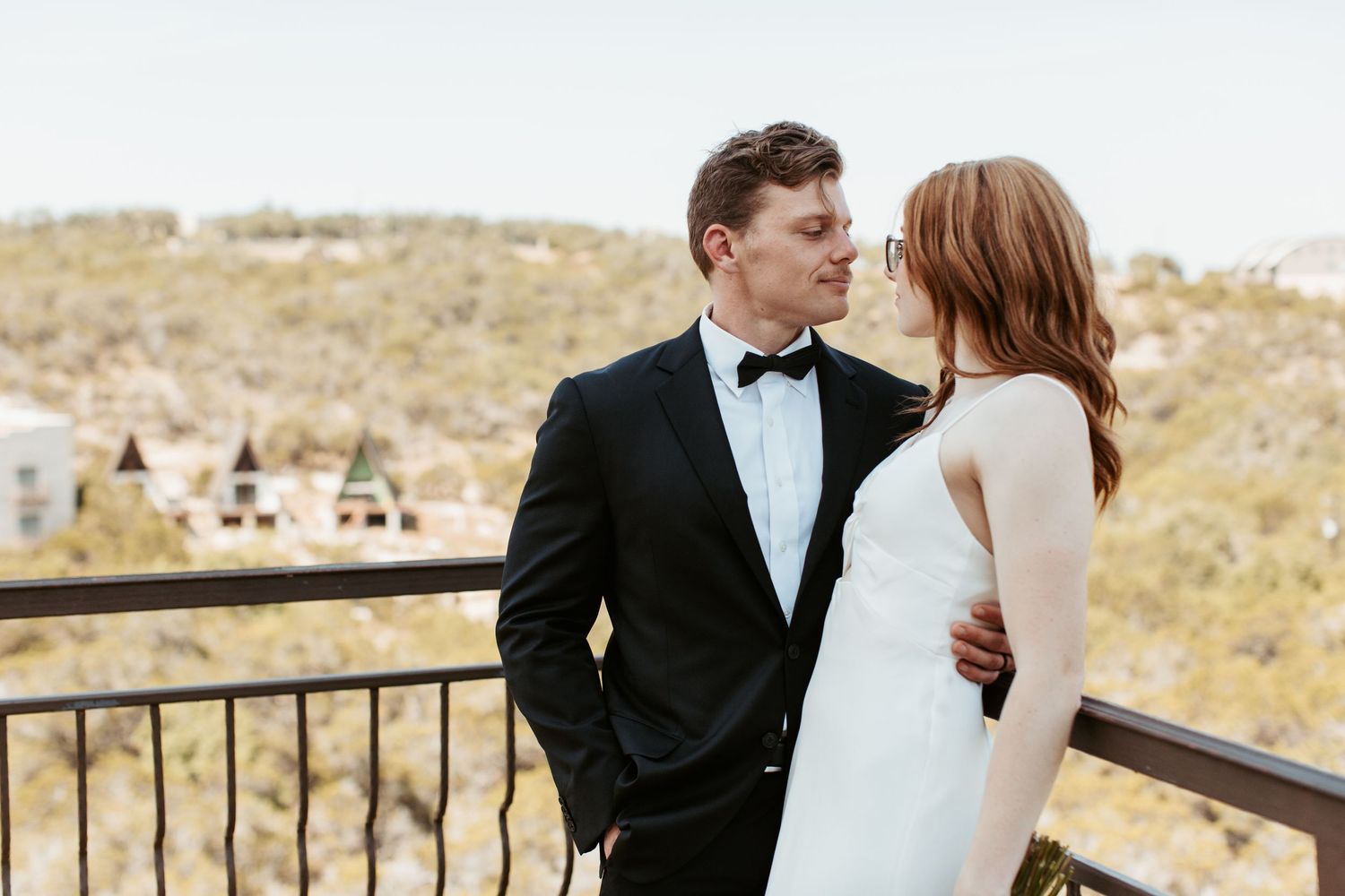 Couple in wedding attire sharing a romantic moment on a balcony with scenic hills in the background.